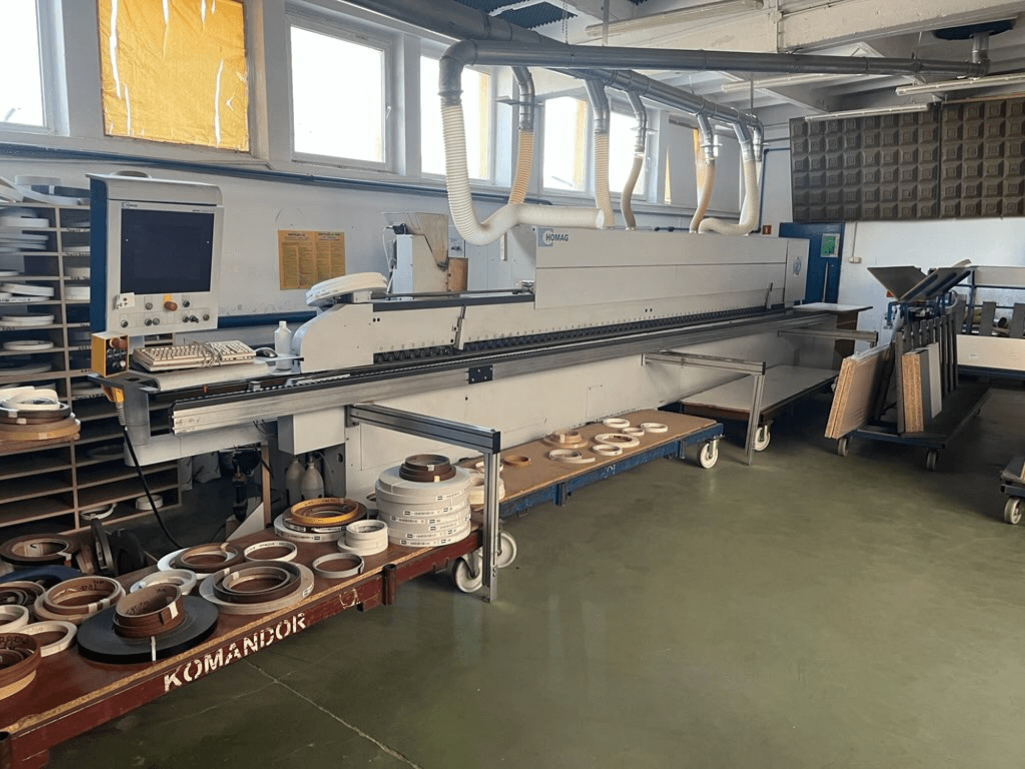 Plaqueuse de chants HOMAG dans un atelier, avec des commandes et divers matériaux de placage de chants sur un chariot bleu. Vue de côté.