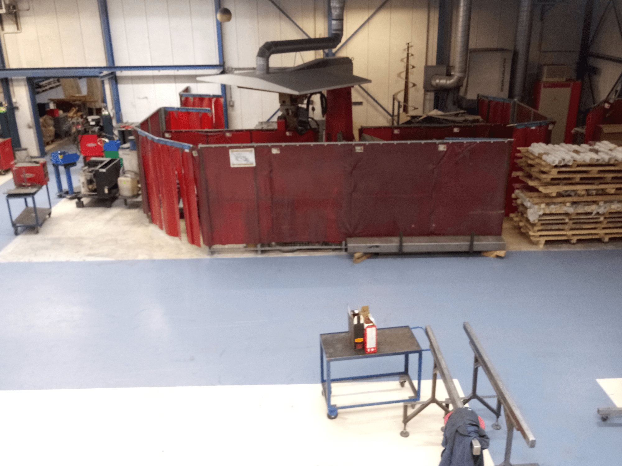 Espace de travail industriel comprenant des rideaux de soudure rouges, des machines et des palettes ; vue aérienne montrant l'agencement et l'équipement.