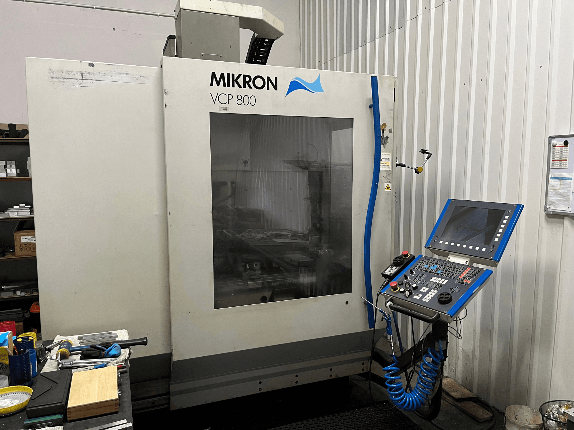 Centre d'usinage vertical MIKRON VCP 800, vue de face, avec le panneau de commande et les outils posés sur un établi au premier plan.
