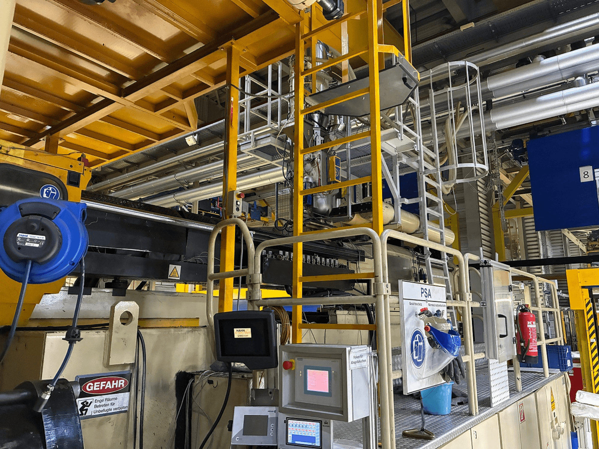 Panneau de commande d'une machine industrielle avec équipement de sécurité bleu et échafaudage jaune, vue latérale montrant les tuyaux et les panneaux d'avertissement.