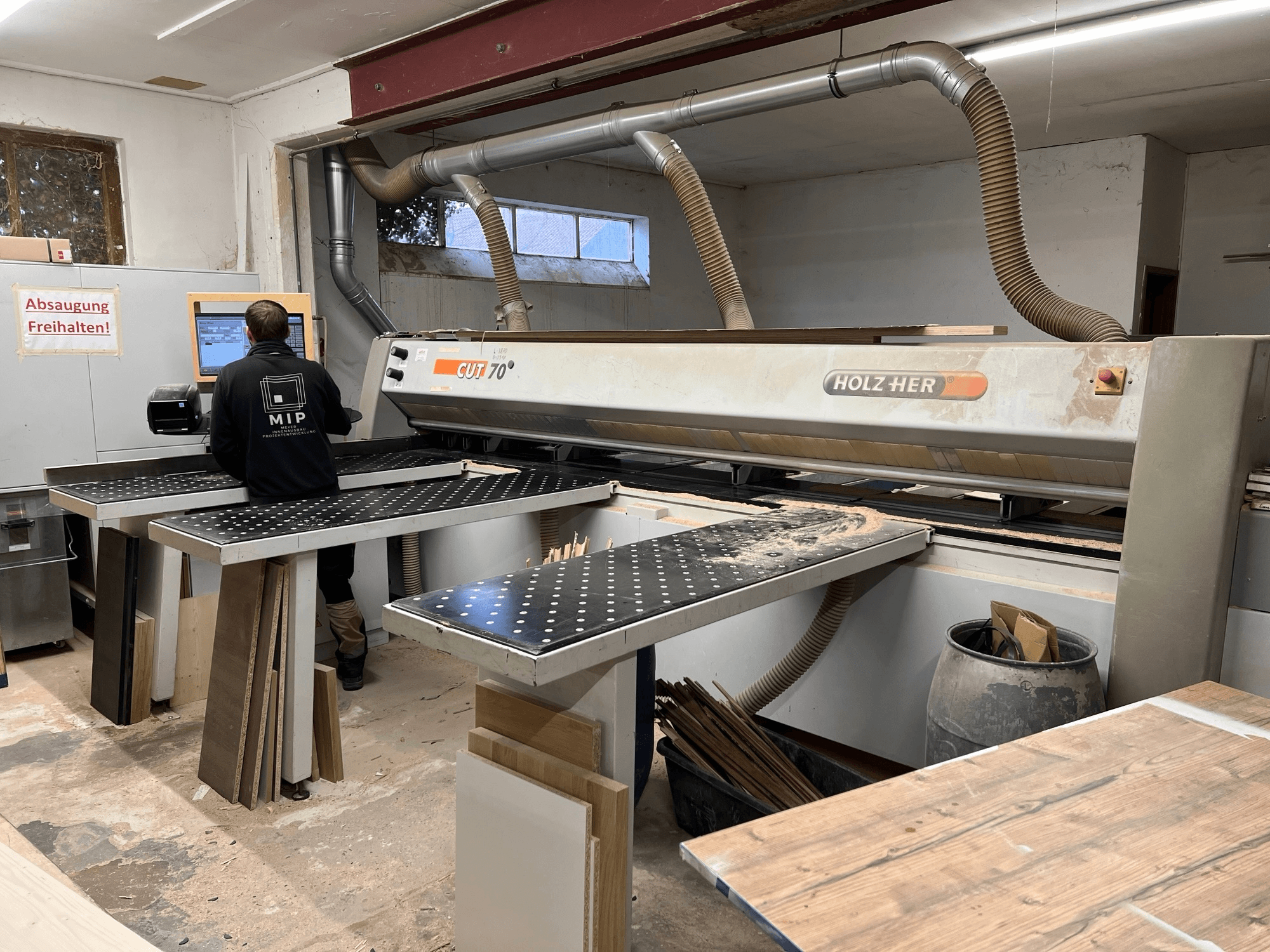 Machine à découper HOLZHER CUT 70 dans un atelier ; vue de face montrant l'opérateur, les tables de travail et les matériaux en bois empilés sur les surfaces.