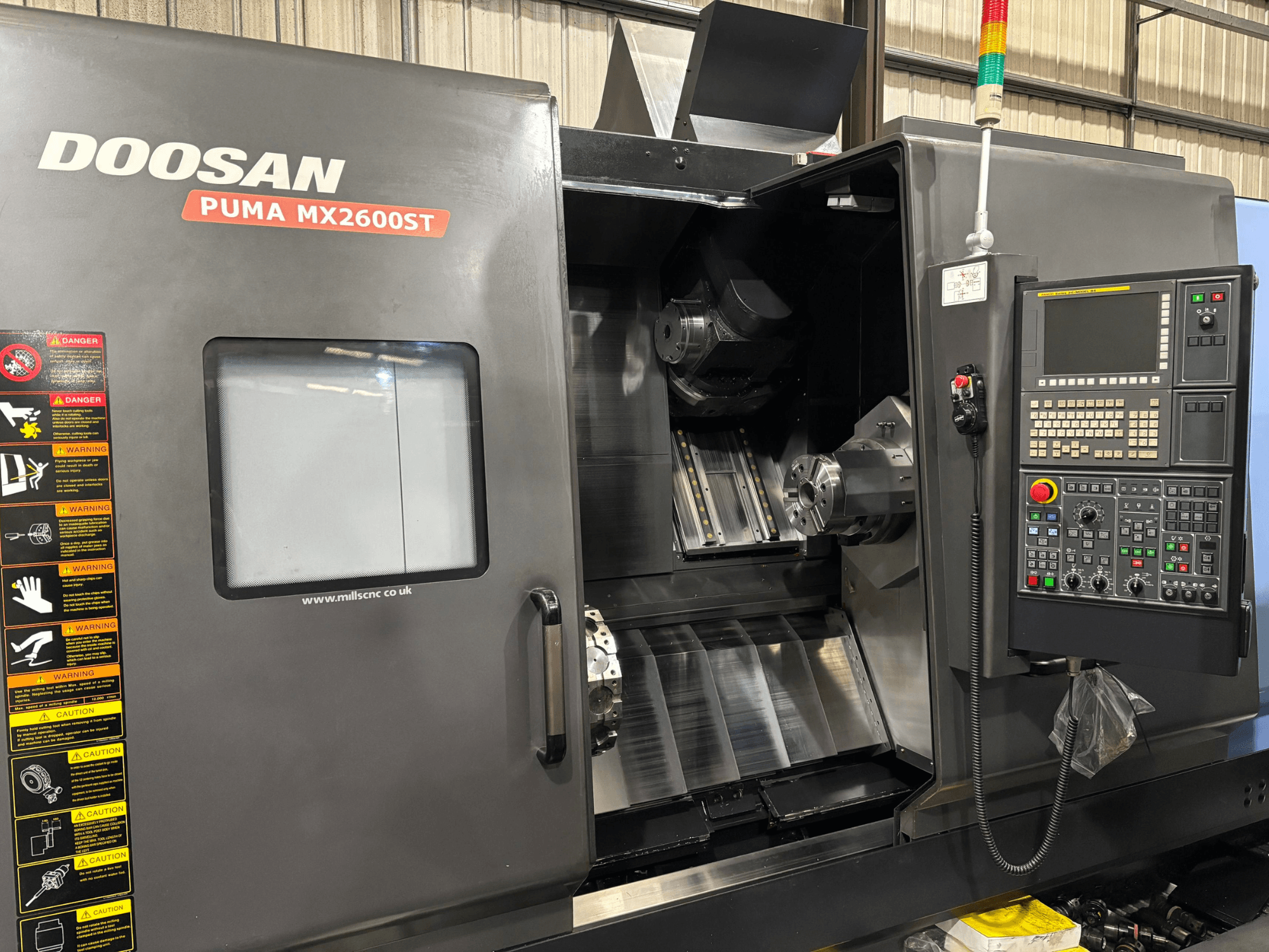 Tour CNC Doosan PUMA MX2600ST, vue de face, montrant le panneau de commande, la broche et la zone d'usinage avec les avertissements de sécurité visibles.