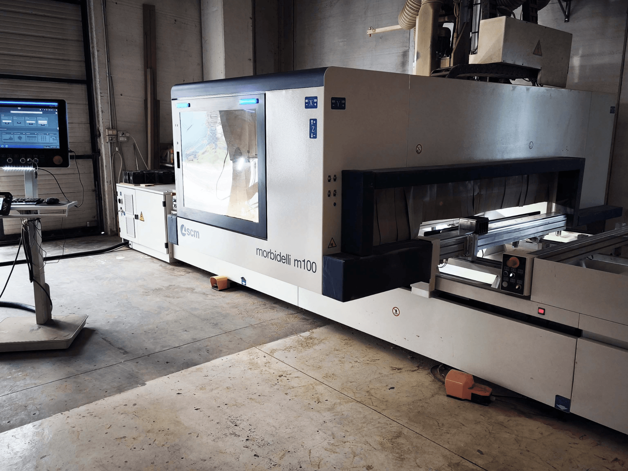 Centre d'usinage CNC SCM Morbidelli M 100 pour le bois, vu de profil, mettant en évidence le panneau de commande, la zone de travail et les dispositifs de sécurité.