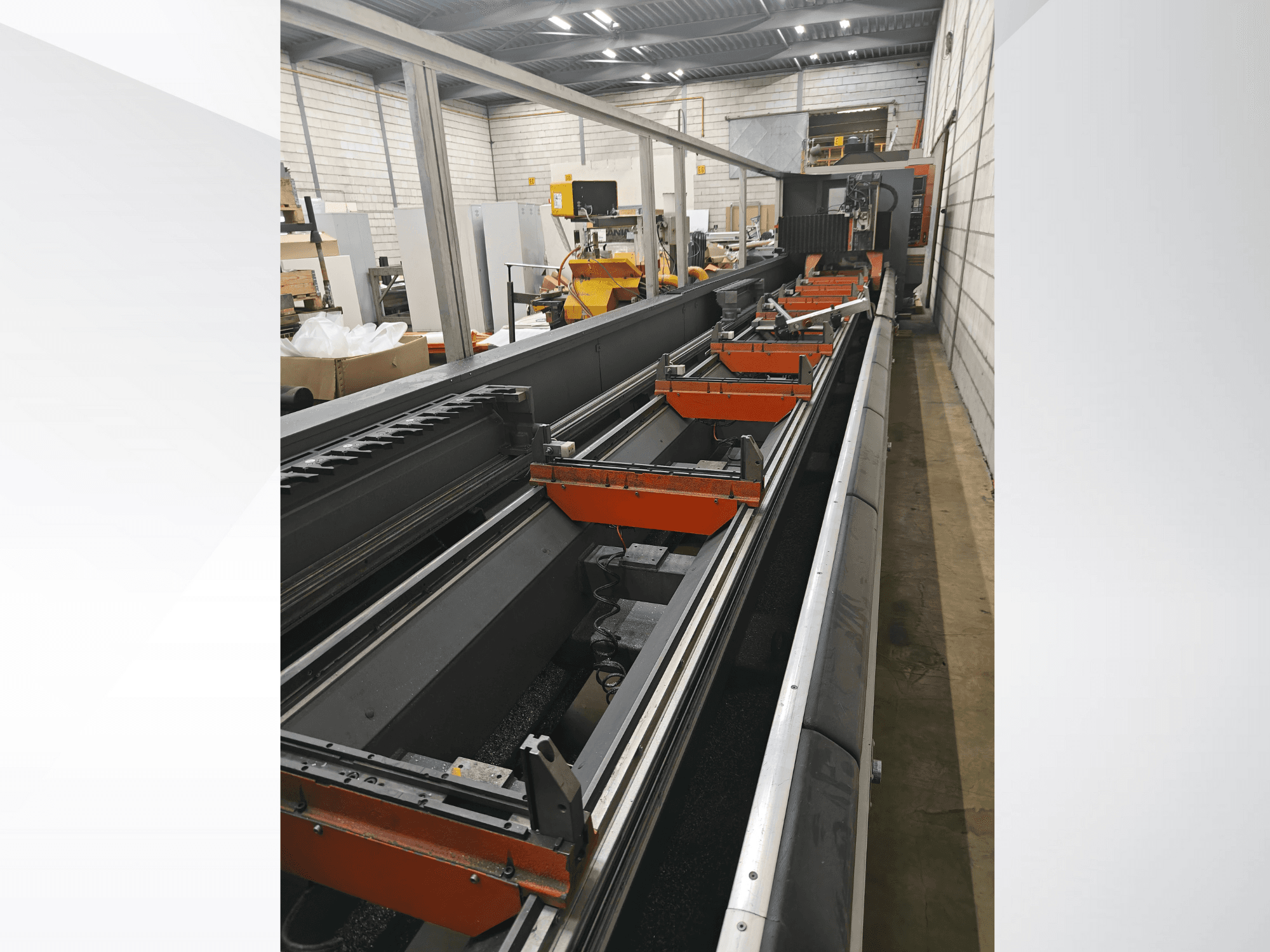 Vue latérale d'une machine industrielle à commande numérique, avec des rails linéaires, des pinces orange et des composants de précision dans un entrepôt.