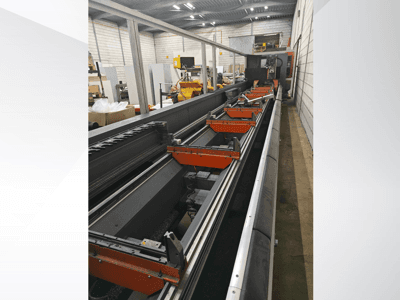 Vue latérale d'une machine industrielle à commande numérique, avec des rails linéaires, des pinces orange et des composants de précision dans un entrepôt.