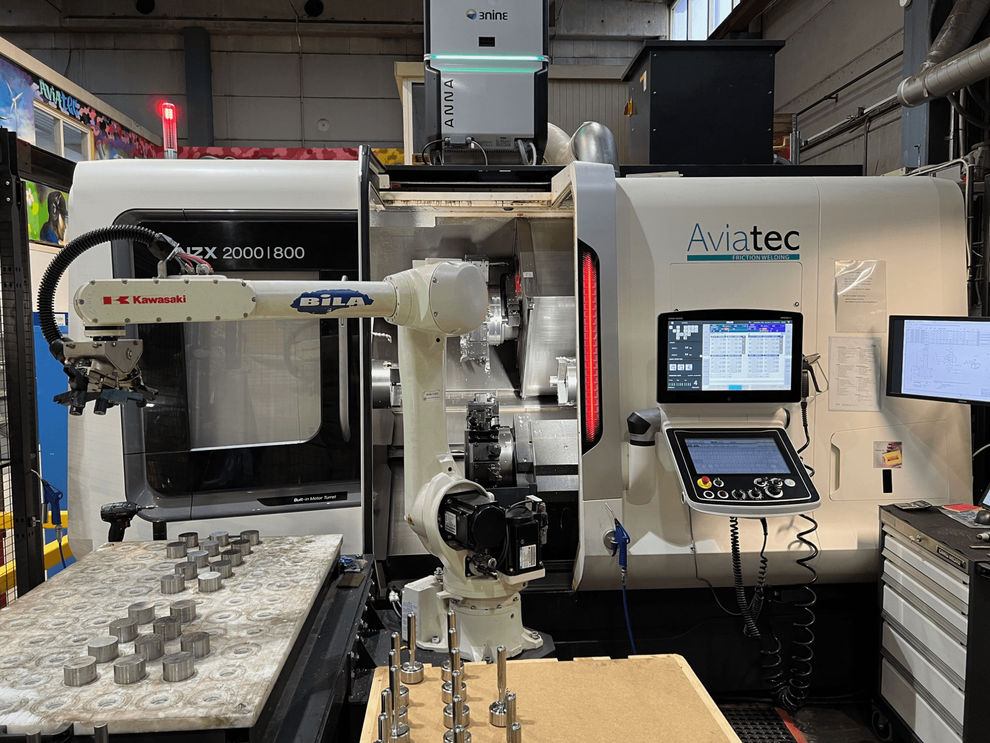 Tour horizontal DMG MORI NZX 2000/800SY équipé d'un bras robotisé Kawasaki, d'un panneau de commande et de pièces métalliques sur la table de travail.