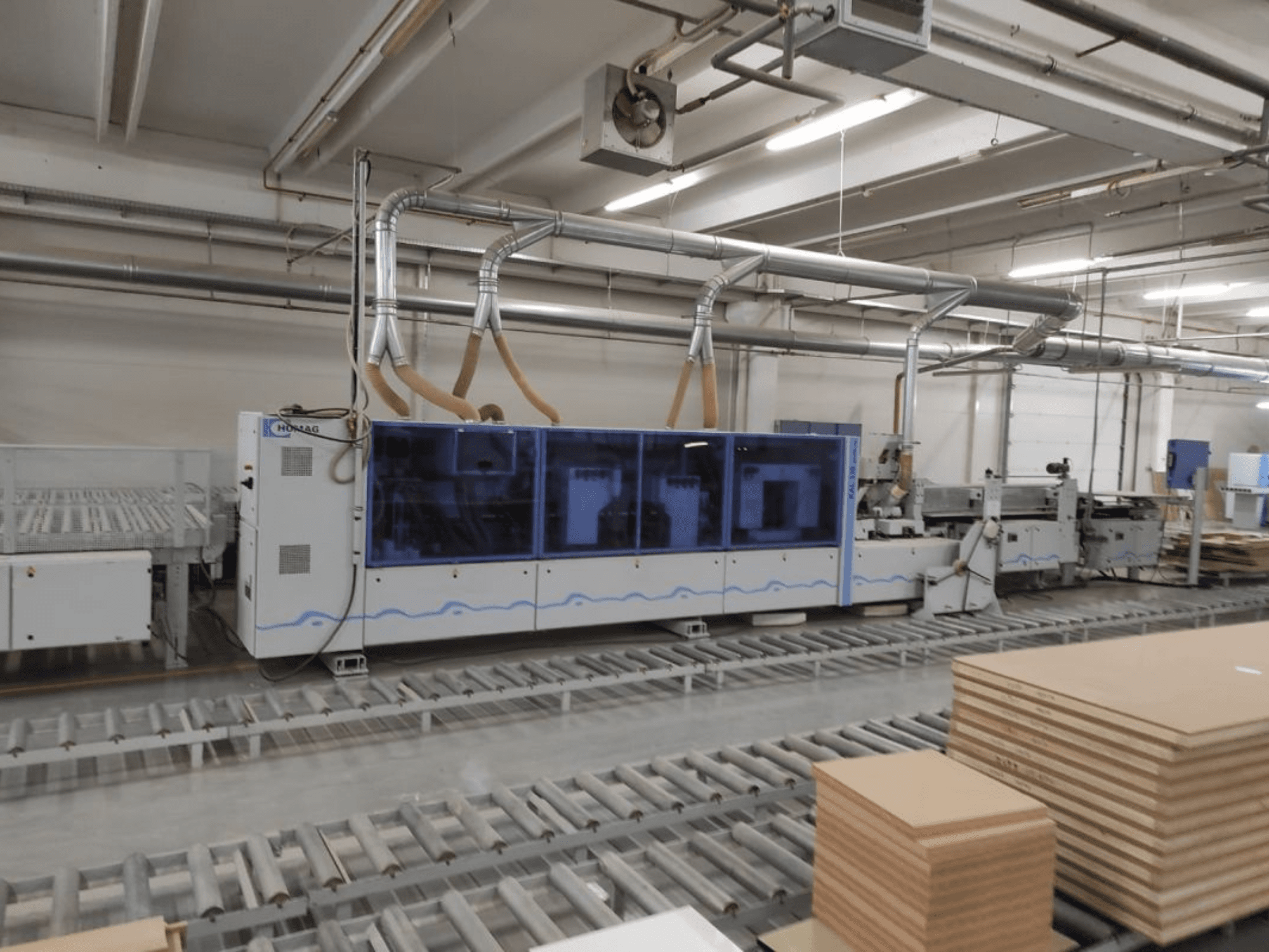 Machine de production HOMAG dans une usine, vue de face, comprenant un boîtier bleu et un système de convoyage avec des feuilles de contreplaqué empilées.