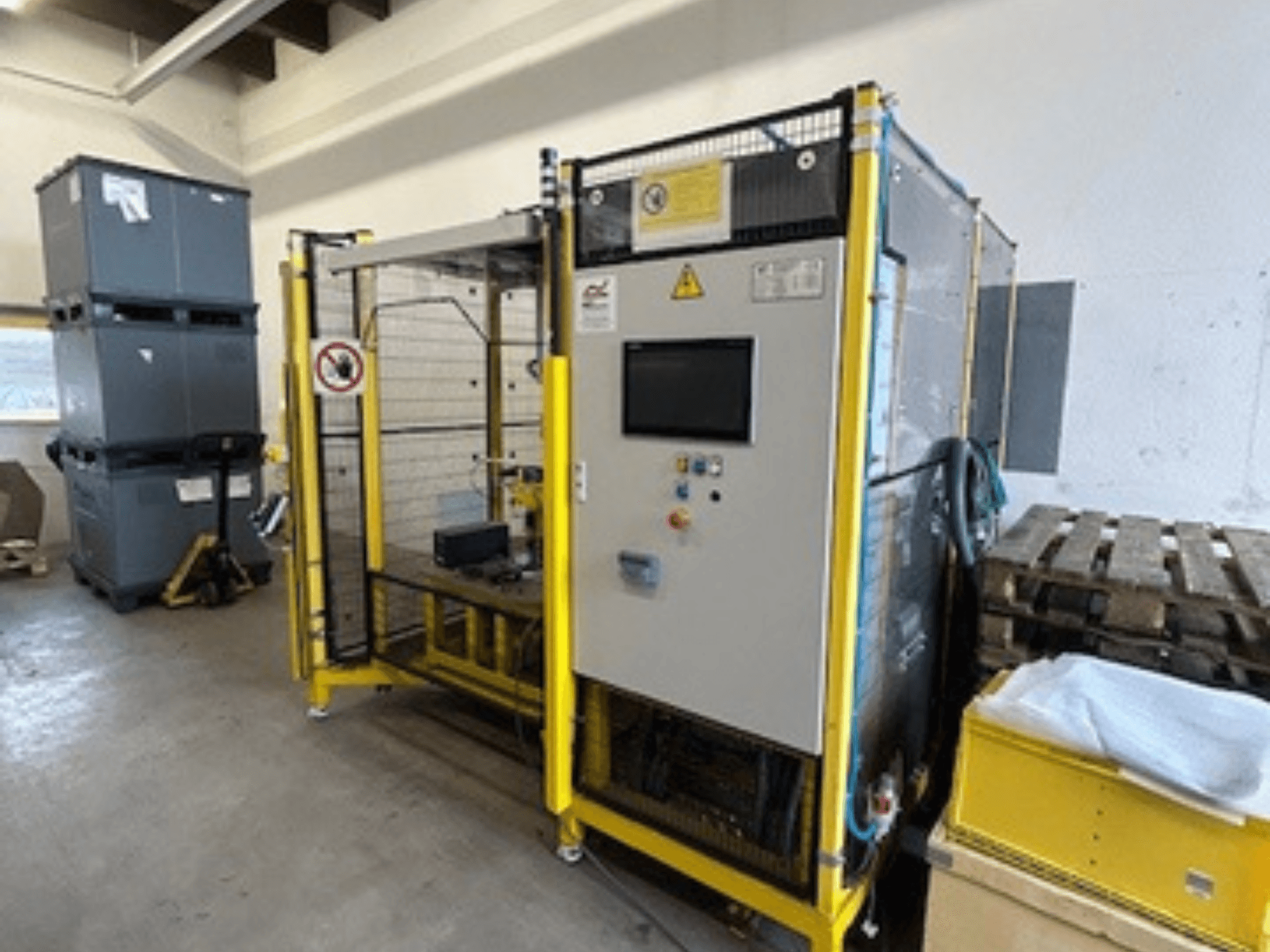 Machine industrielle avec panneau de commande et dispositifs de sécurité, vue de face ; cadre jaune, bacs gris et palettes en arrière-plan.