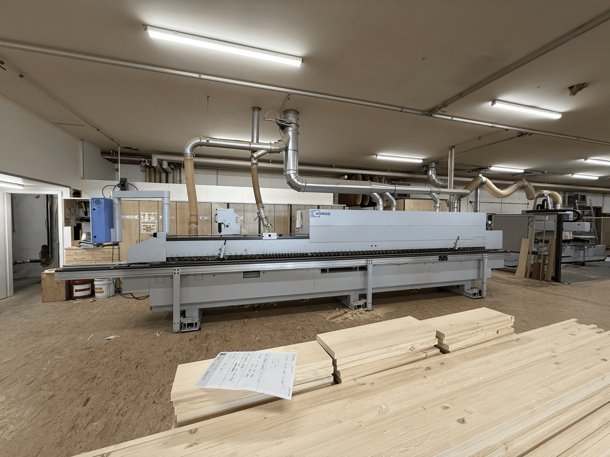 HOMAG KAL211 Plaqueuse de chants dans un atelier, vue de face, avec des planches de bois et des machines dans un environnement bien éclairé.