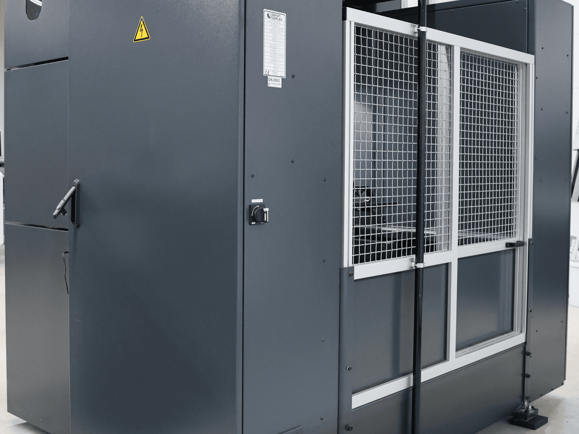 Machine industrielle grise avec avertissement de sécurité, vue de face montrant la porte grillagée et le panneau de commande, modèle étiqueté.