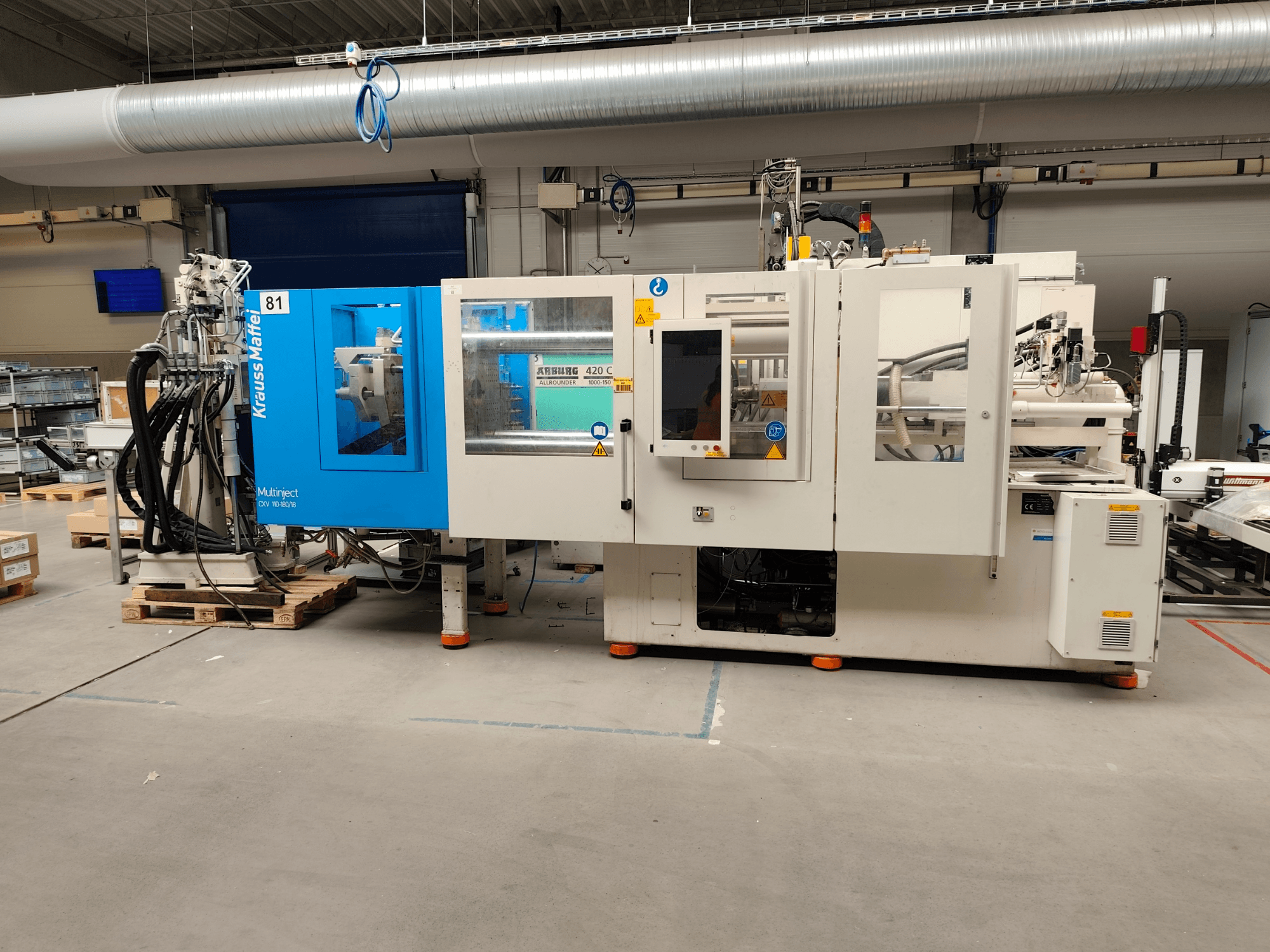 Presse à injection hydraulique Krauss Maffei 110-180-180 CX V, vue de profil, mettant en évidence le châssis bleu et blanc, le panneau de commande et les connecteurs.