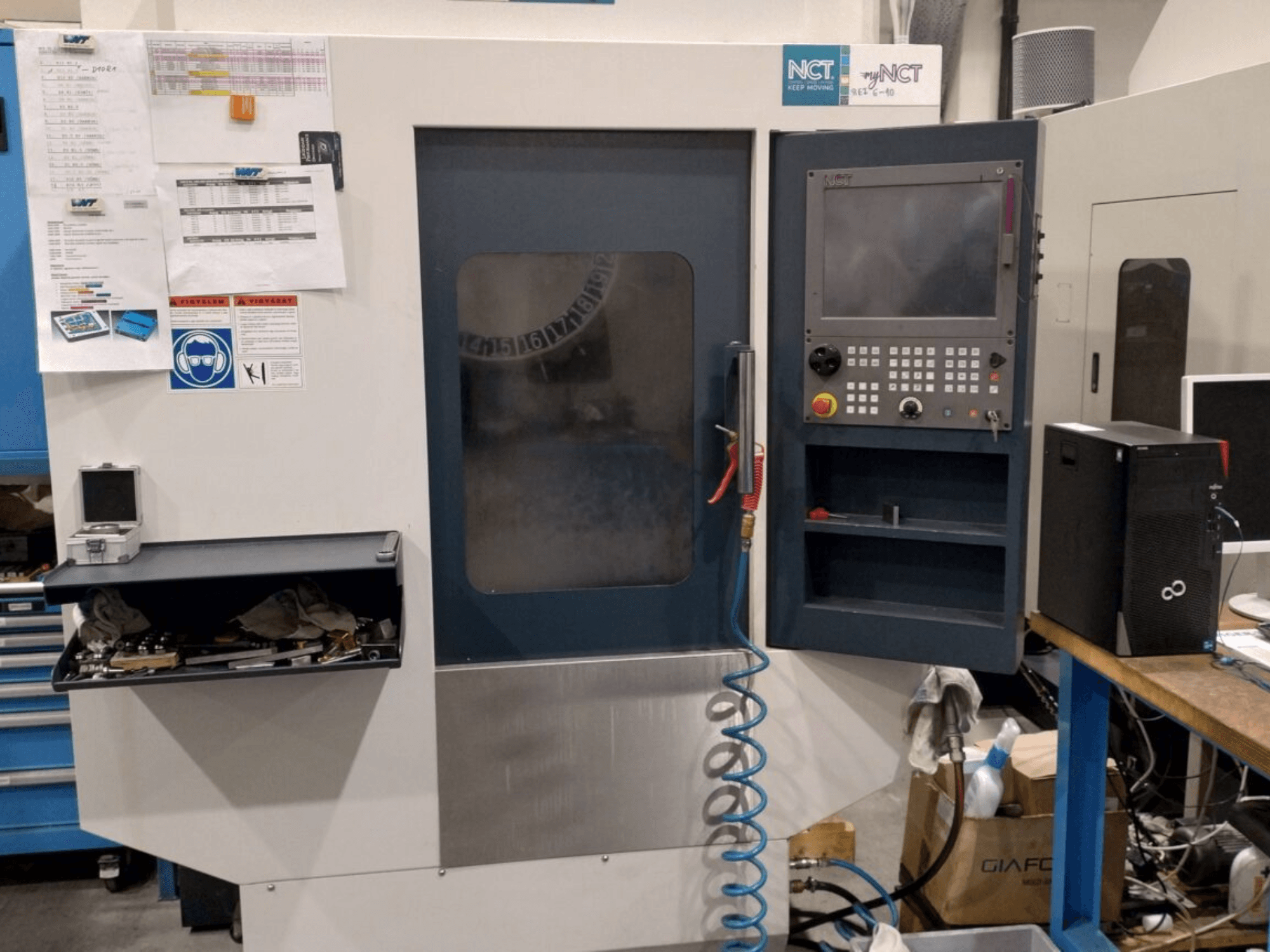 Machine CNC NCT, vue de face, comprenant un panneau de commande avec clavier numérique, des outils dans un tiroir de rangement et un manuel sur le mur.