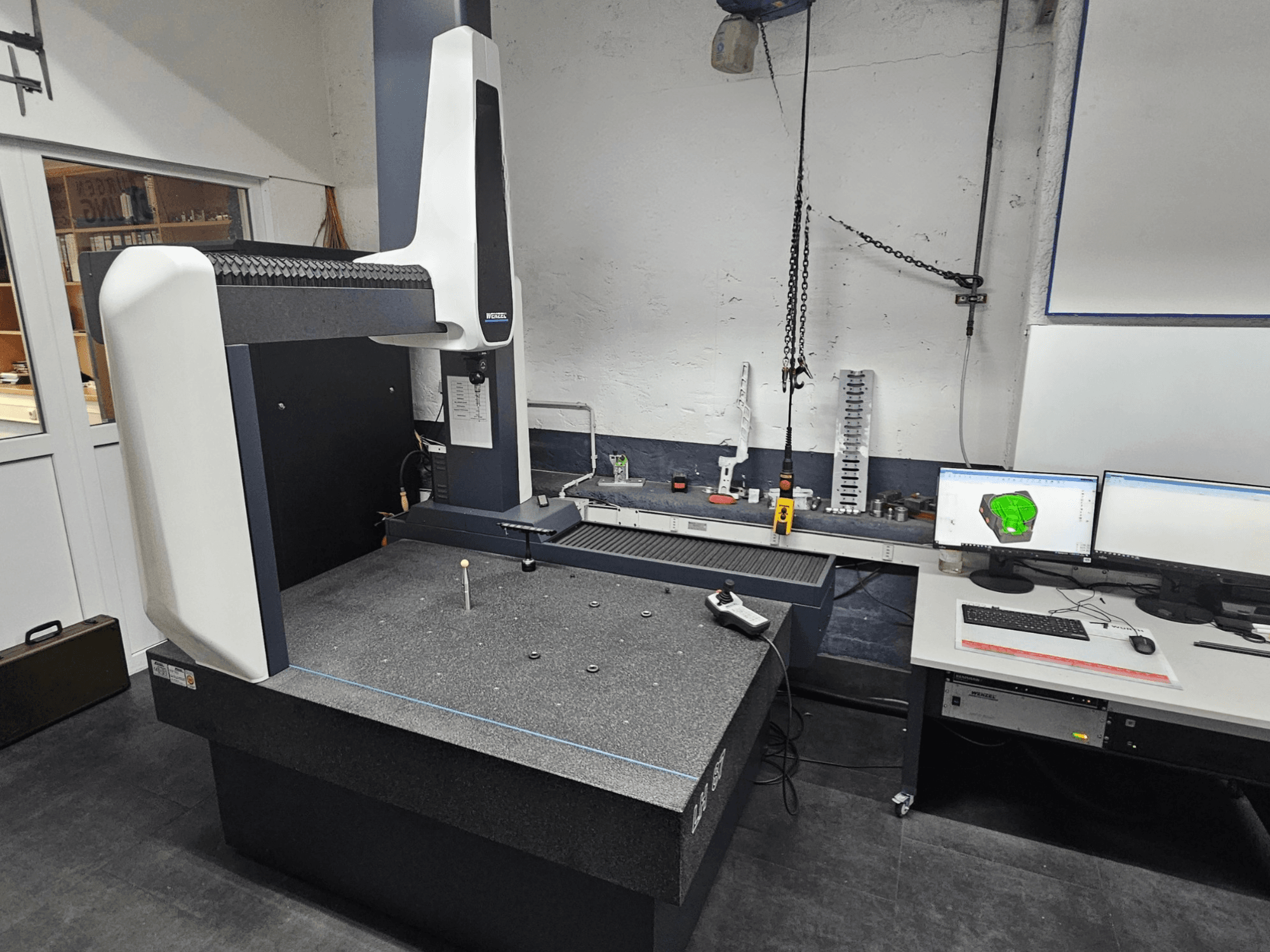 Machine à mesurer tridimensionnelle Wenzel LH 87 dans un atelier, vue de face, avec un palpeur sur la table en granit et deux moniteurs affichant la conception CAO.