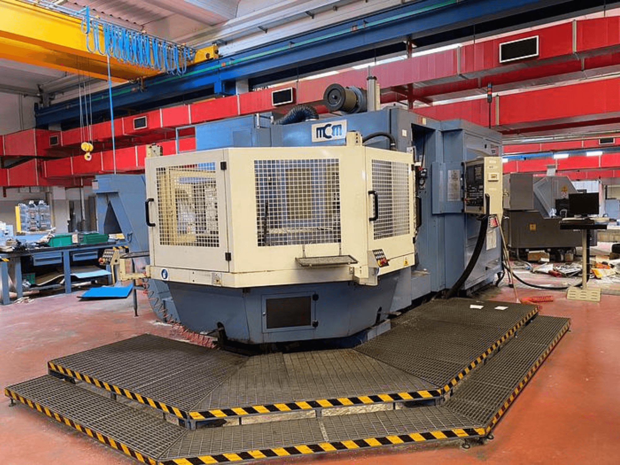 Machine CNC MCM dans une usine, vue de face avec cage de protection, bords de sécurité noirs et jaunes, entourée d'équipements industriels.