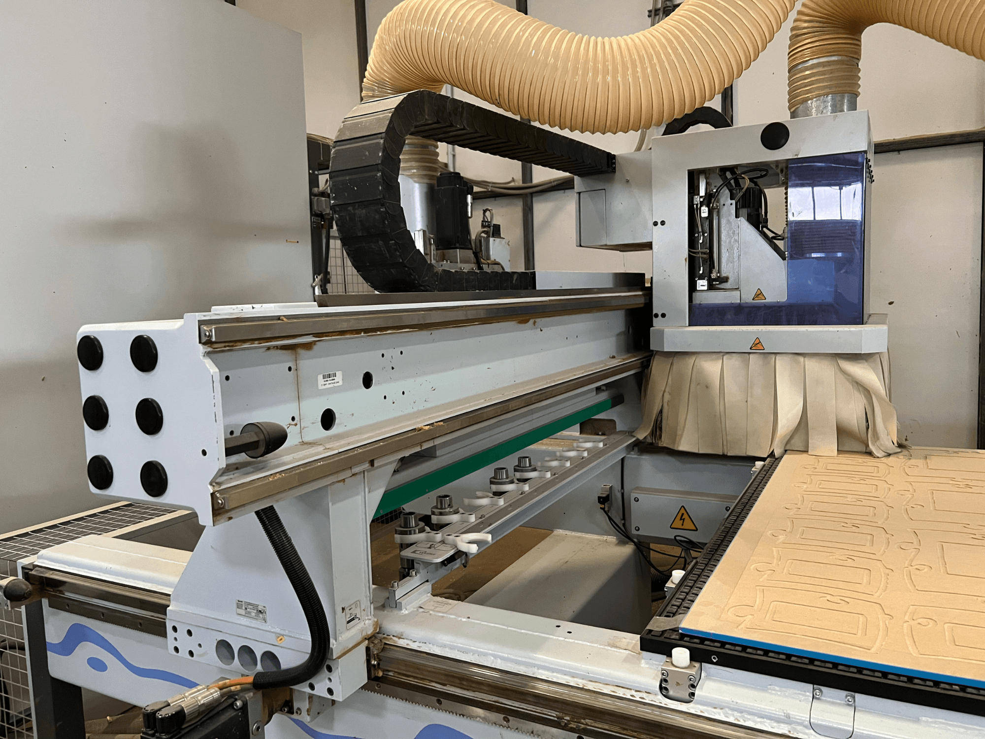 Vue latérale d'une défonceuse CNC avec un panneau de commande bleu et argenté, équipée d'un système d'aspiration des poussières et d'un banc de découpe du bois.