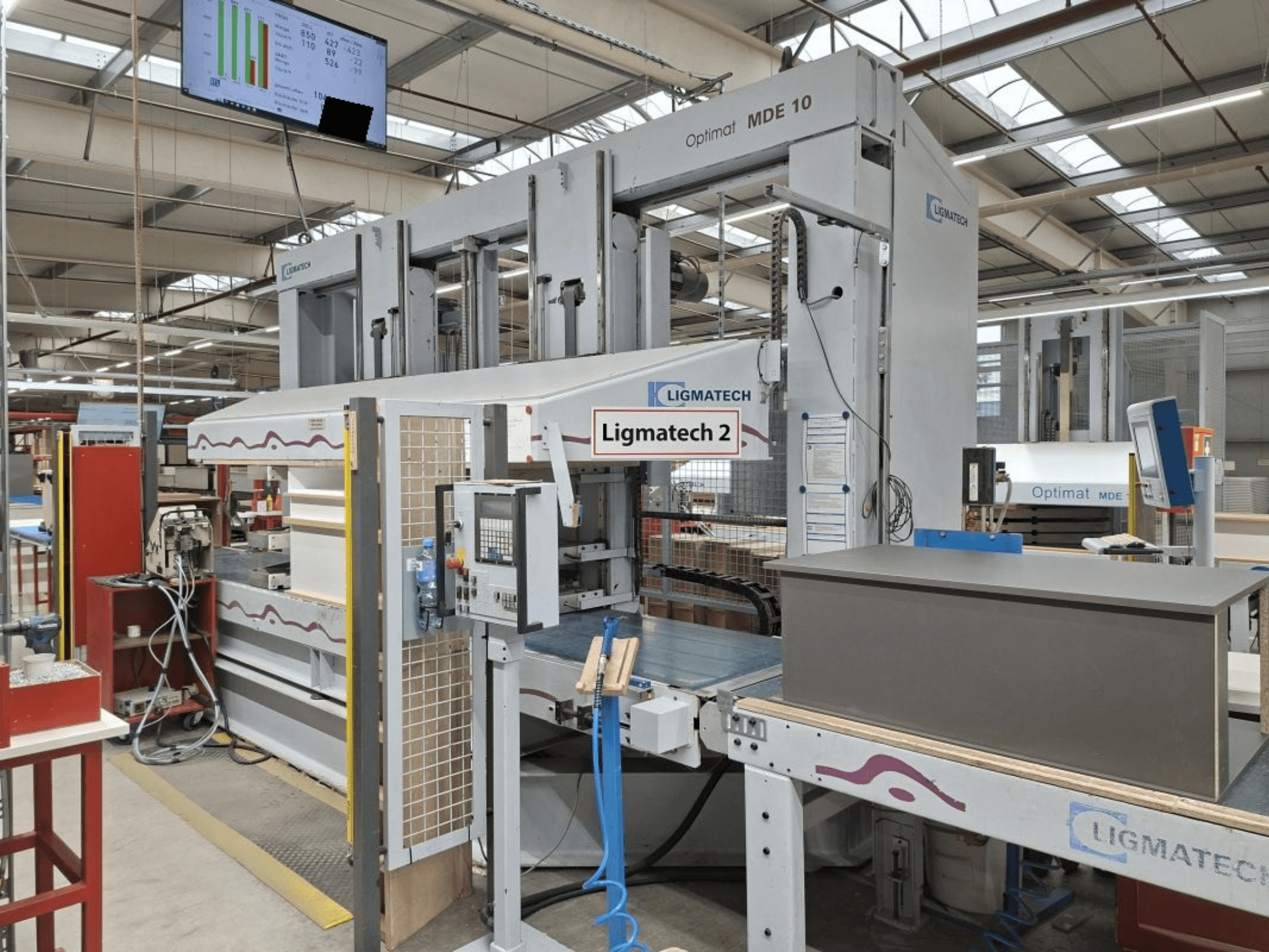 Machine LIGMATECH Optimat MDE 10 dans un atelier, vue latérale montrant les commandes et la pièce de bois sur le convoyeur.