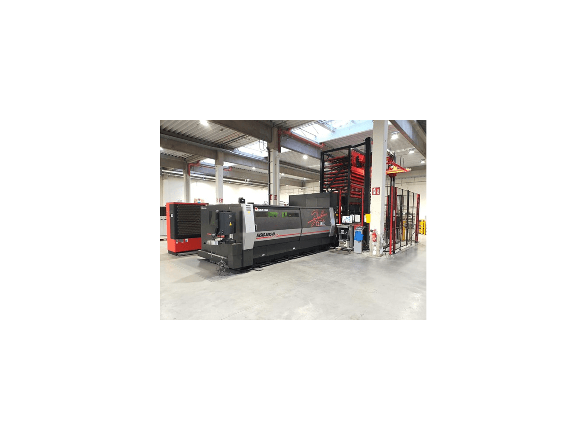 Machine de découpe laser Amada ENSIS 3015 Aj vue de face, installée dans un atelier industriel spacieux avec étagères métalliques.
