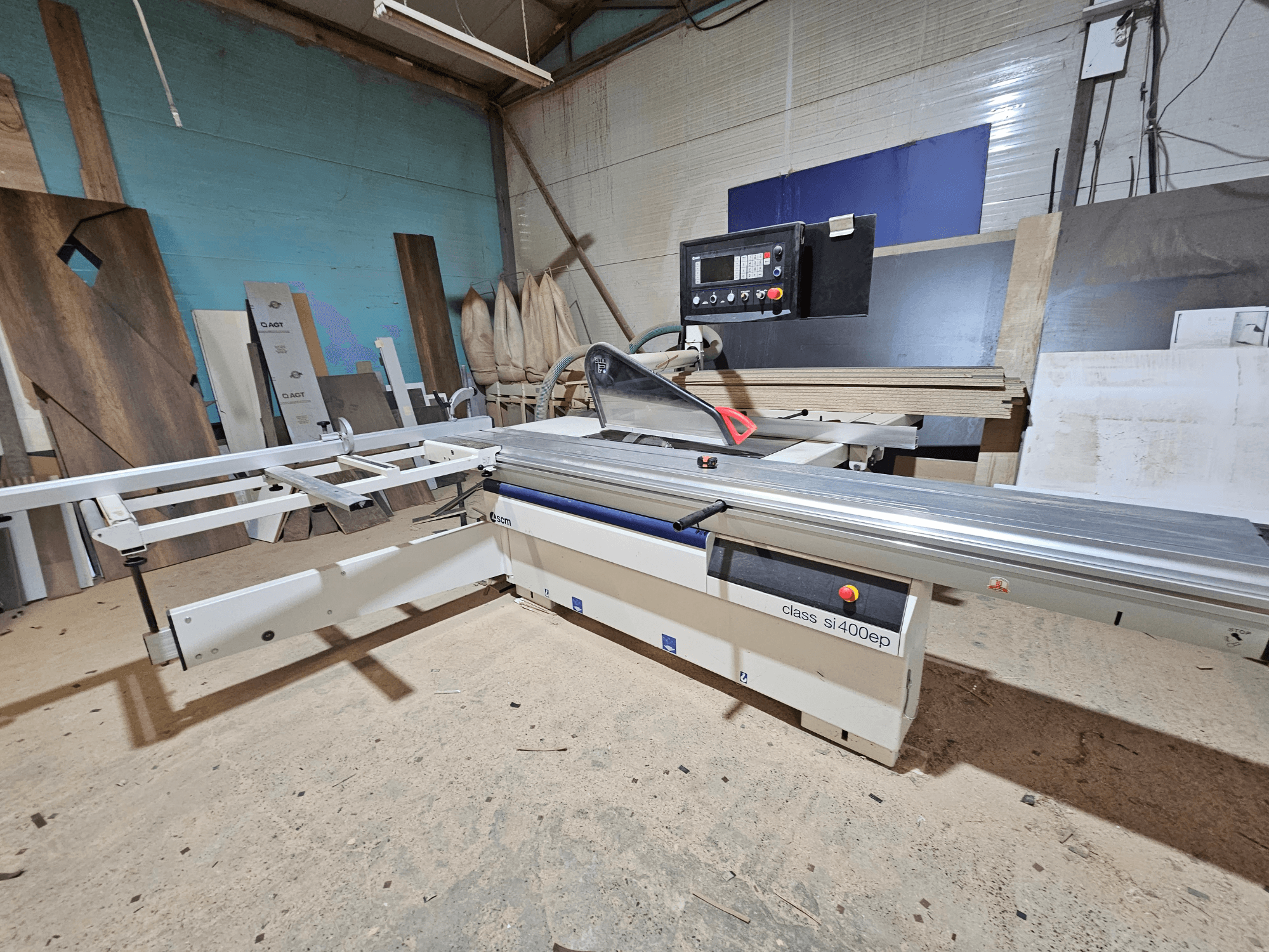 Scie à panneaux SI400EP, vue de face, avec guide réglable, posée sur le sol sablonneux de l'atelier, entourée de panneaux de bois.