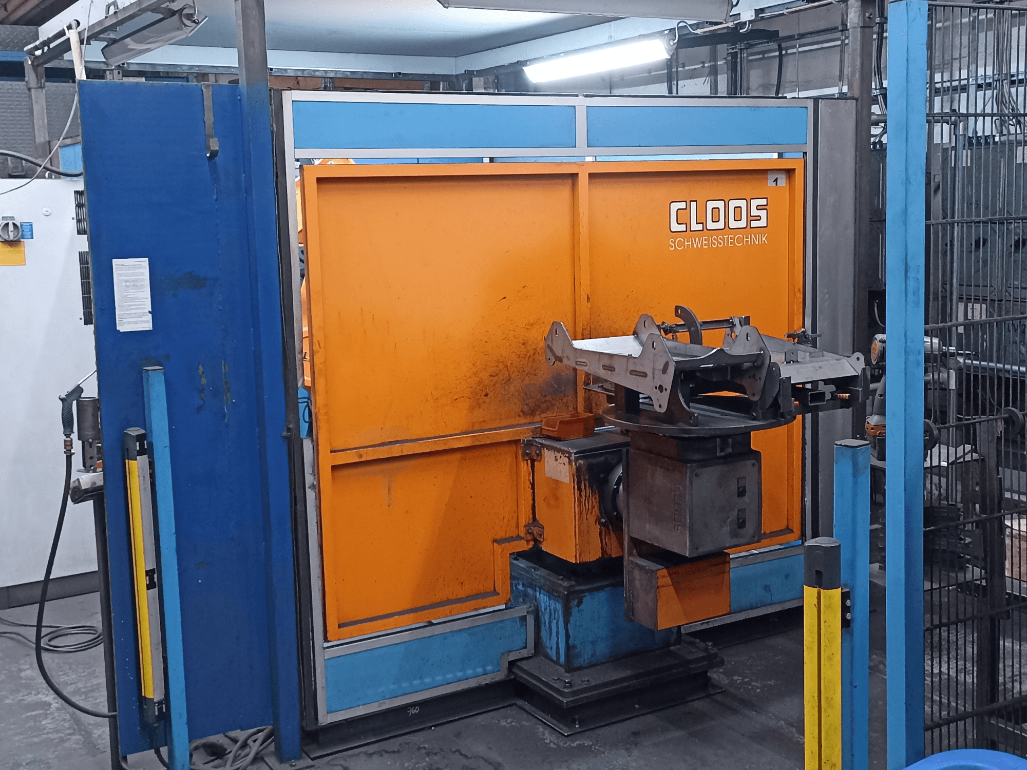 Robot de soudage CLOOS Romat 320 dans un atelier, vue de face, avec un panneau de commande orange et une pièce métallique posée sur un socle rotatif.