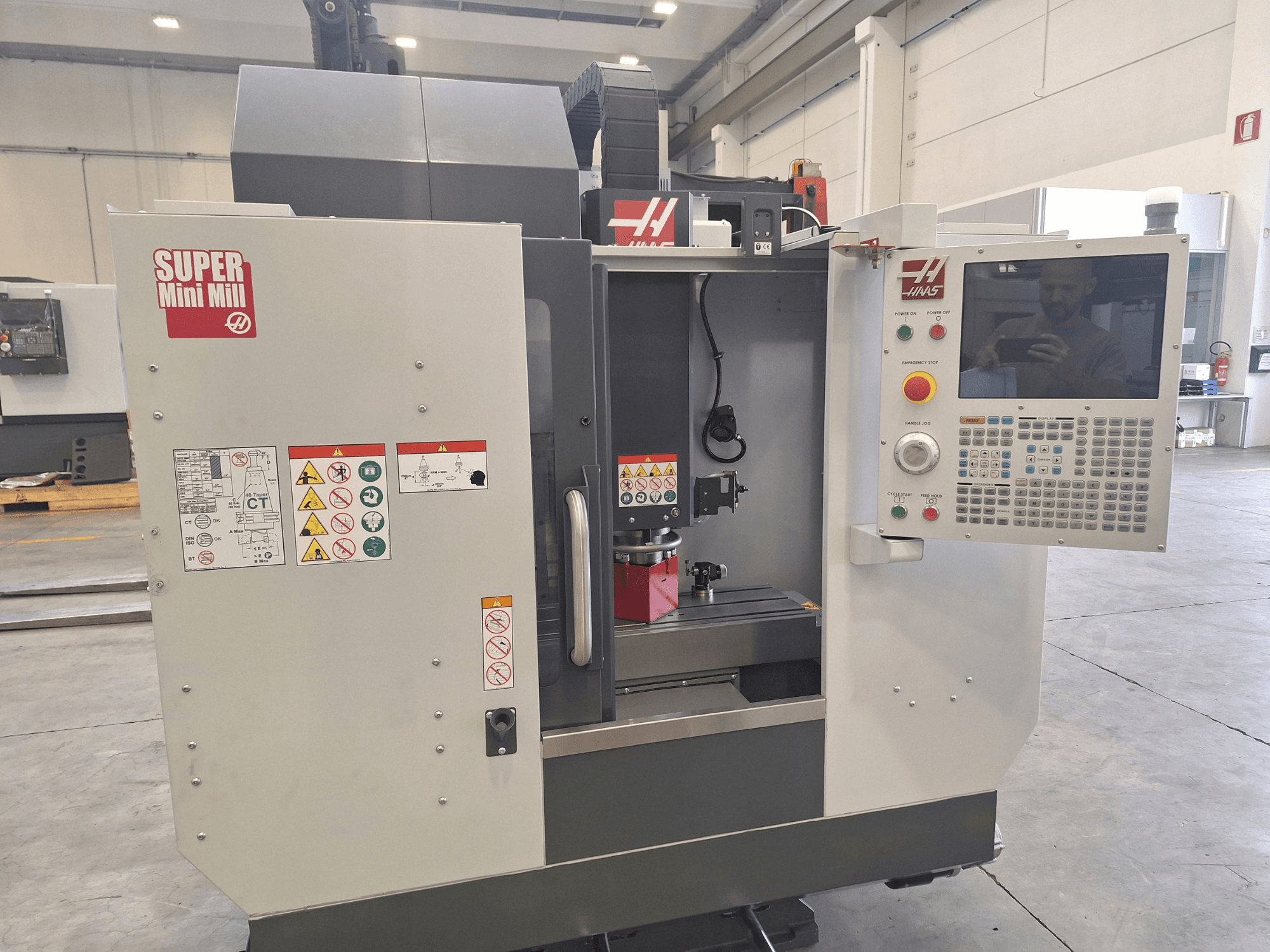 Vue de face de la Super Mini Mill de HAAS montrant le panneau de commande, les étiquettes de sécurité et la zone d'usinage avec une pièce en cours de montage.