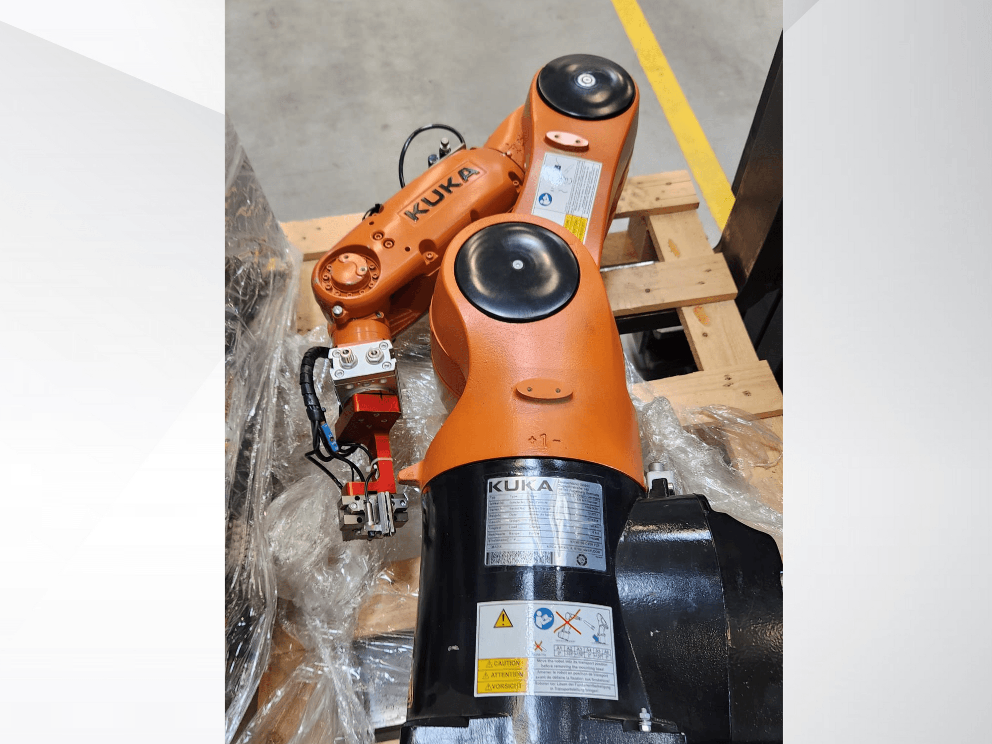 Bras robotique KUKA avec finition orange, vue latérale, comprenant une pince et le câblage de commande sur une palette en bois.