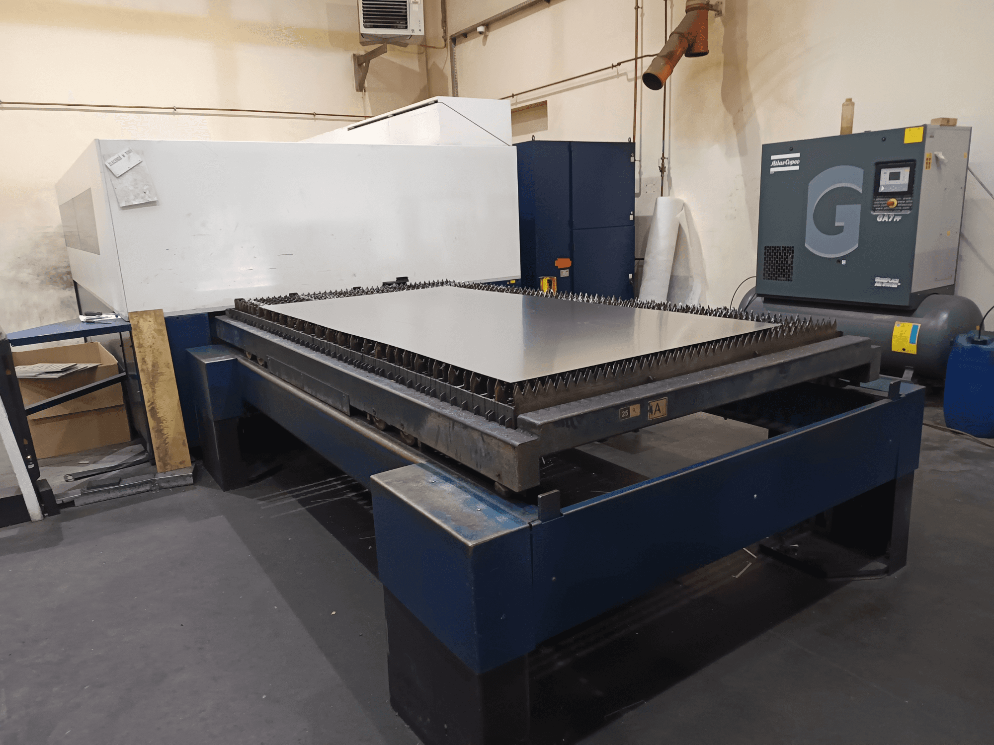 Machine à découper industrielle avec table en métal, vue de face. Elle présente des arêtes vives, un compresseur d'air à droite et un châssis en acier.