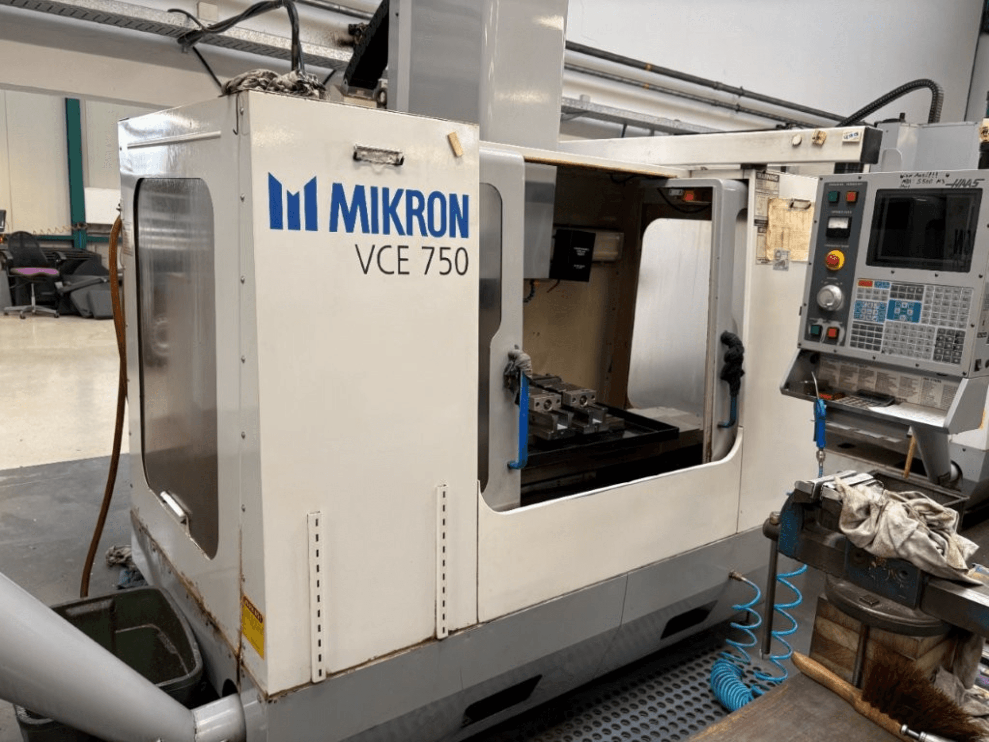 Vue de face de la fraiseuse CNC Mikron VCE 750, avec une table de travail usinée et un panneau de commande avec divers boutons et écrans.