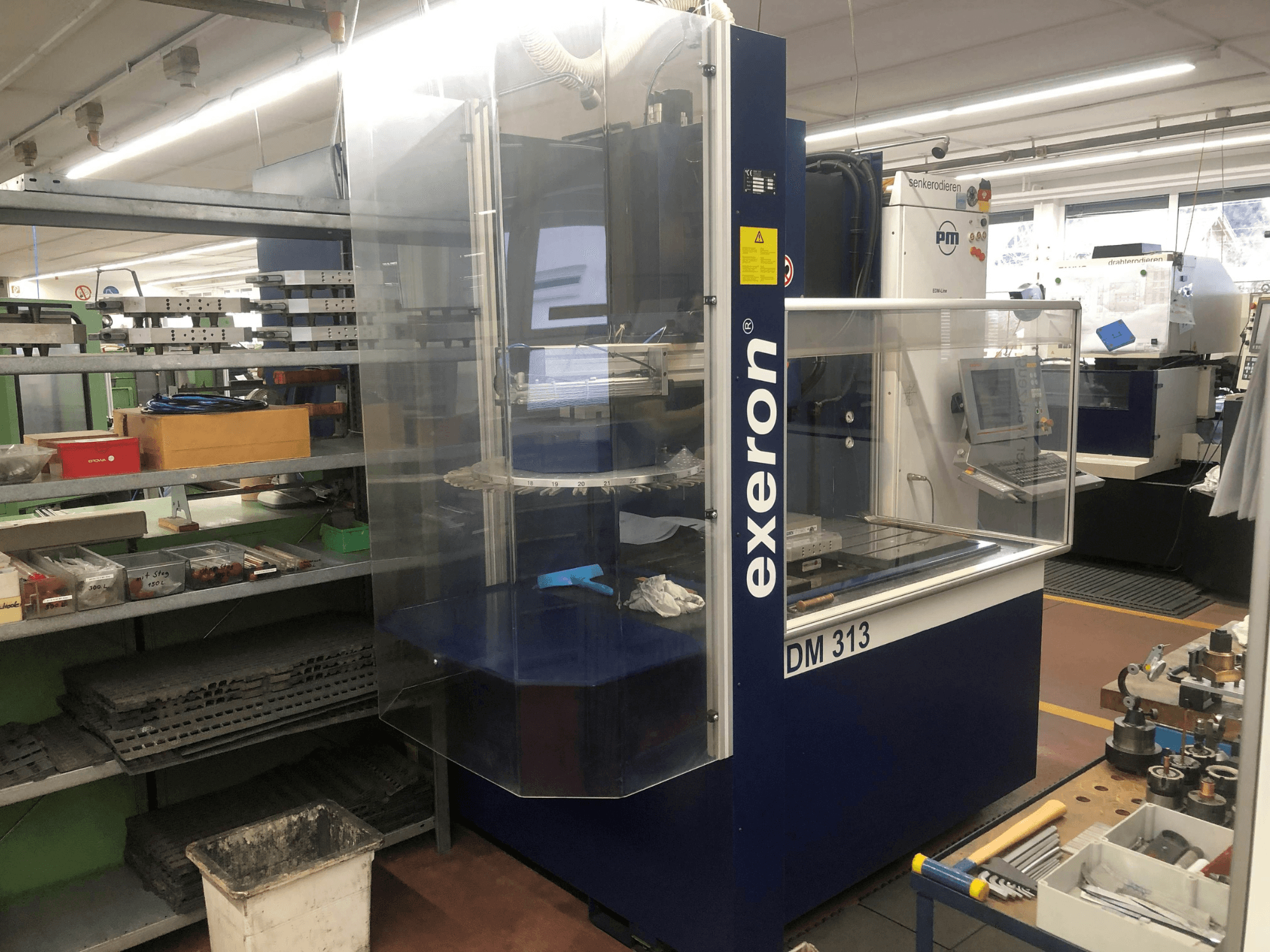 Machine d'enfonçage Exeron EDM 313 dans un atelier, montrant une vue de face avec un boîtier transparent et un panneau de commande.