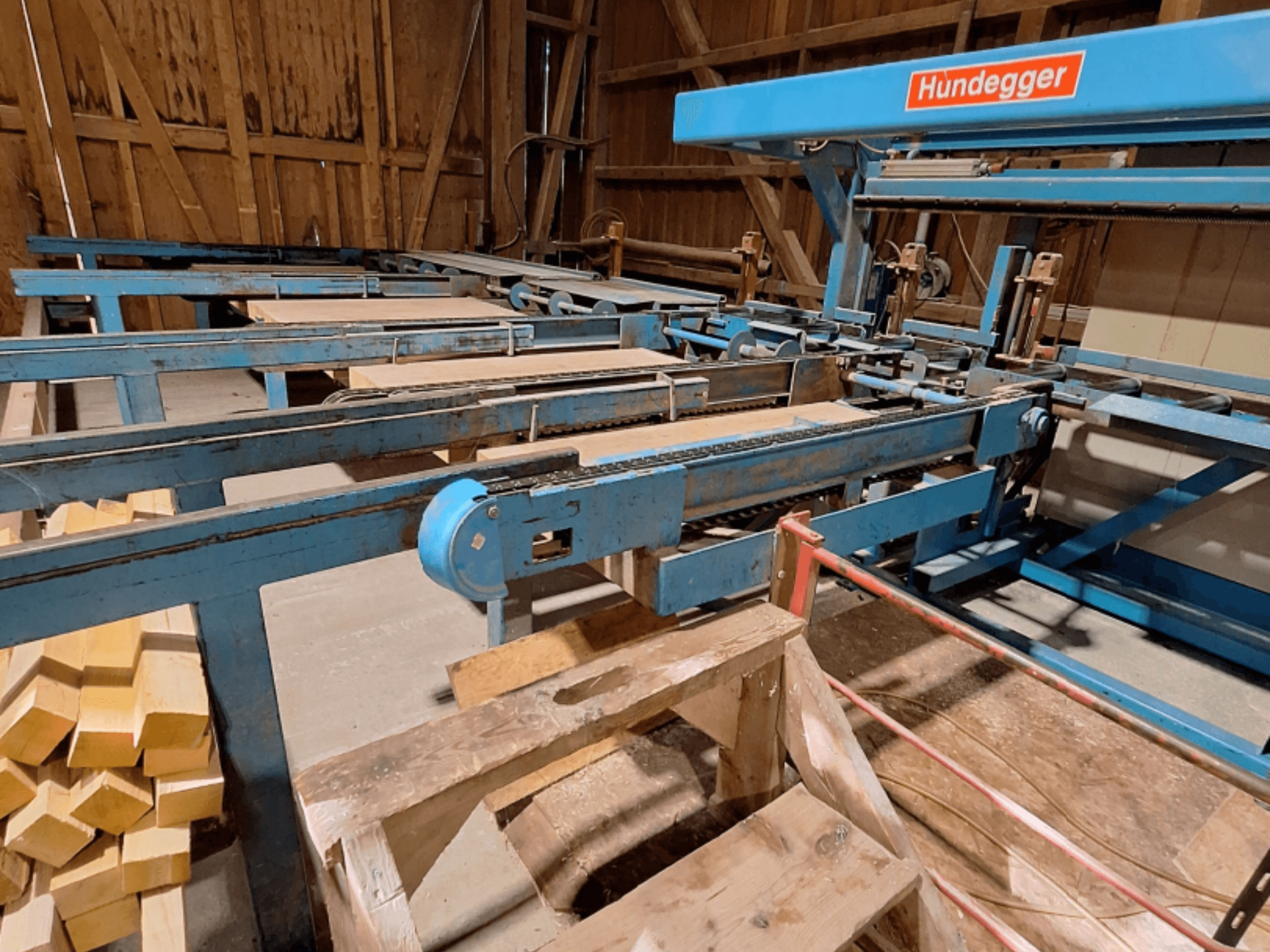 Centre d'usinage CNC du bois HUNDEGGER K2-A installé dans une grange, avec son châssis métallique bleu, son système de convoyage et ses poutres en bois empilées.