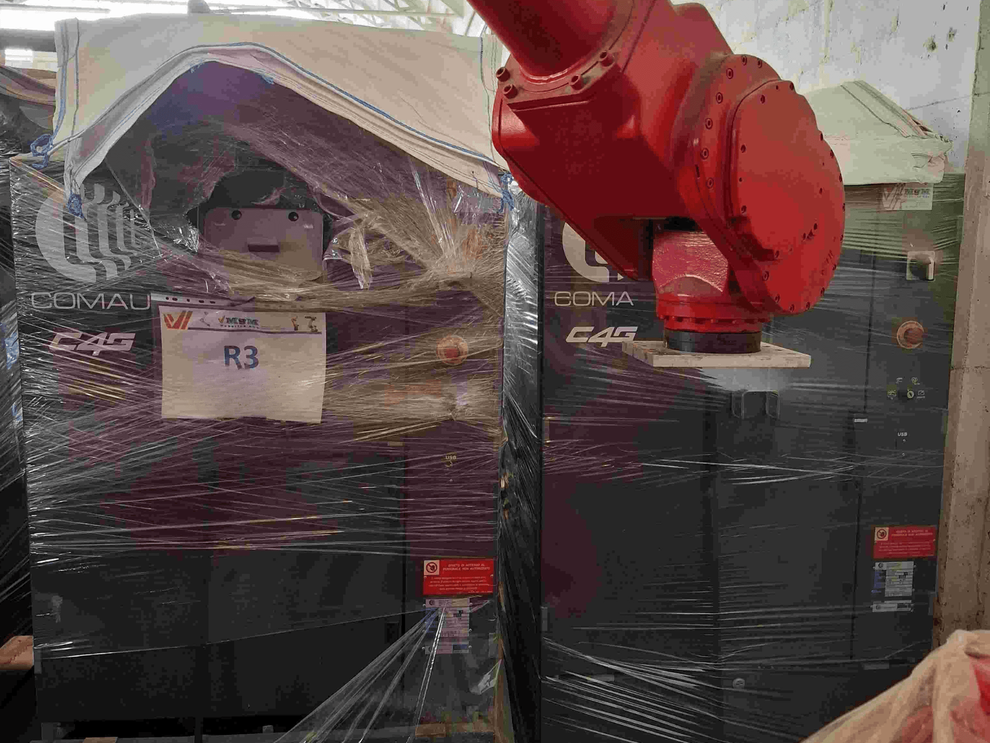 Bras robotique C4G de Comau vu de face, emballé dans du plastique, étiqueté "R3", avec des autocollants d'avertissement visibles sur la machine.