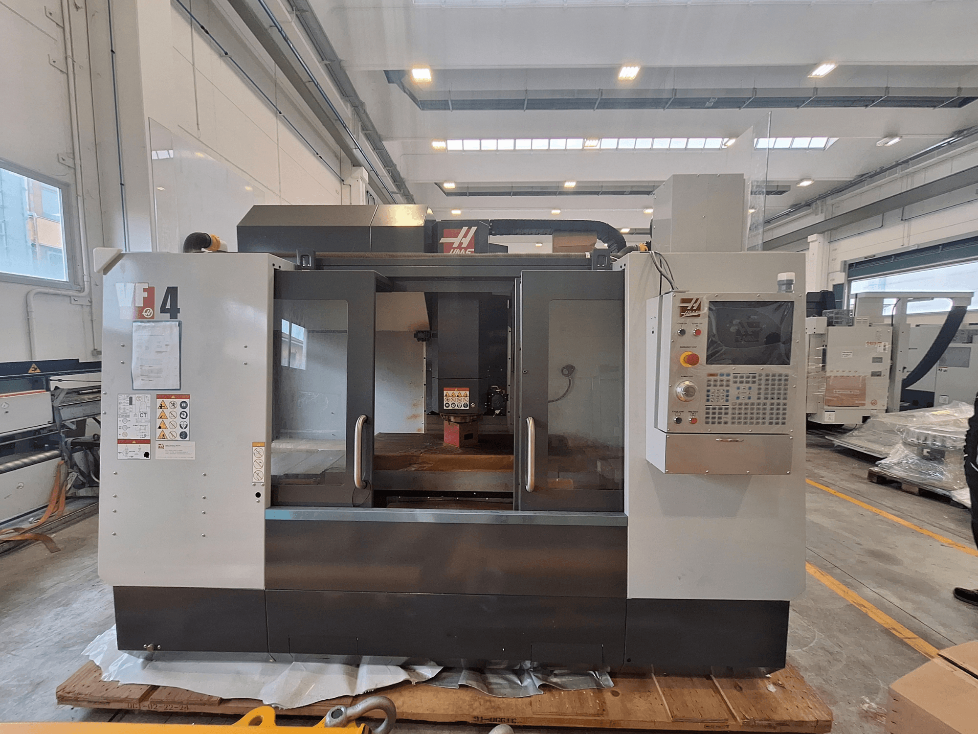 Centre d'usinage vertical HAAS VF-4, vue de face, présentant le panneau de commande, la zone de travail et les étiquettes de sécurité dans un entrepôt.