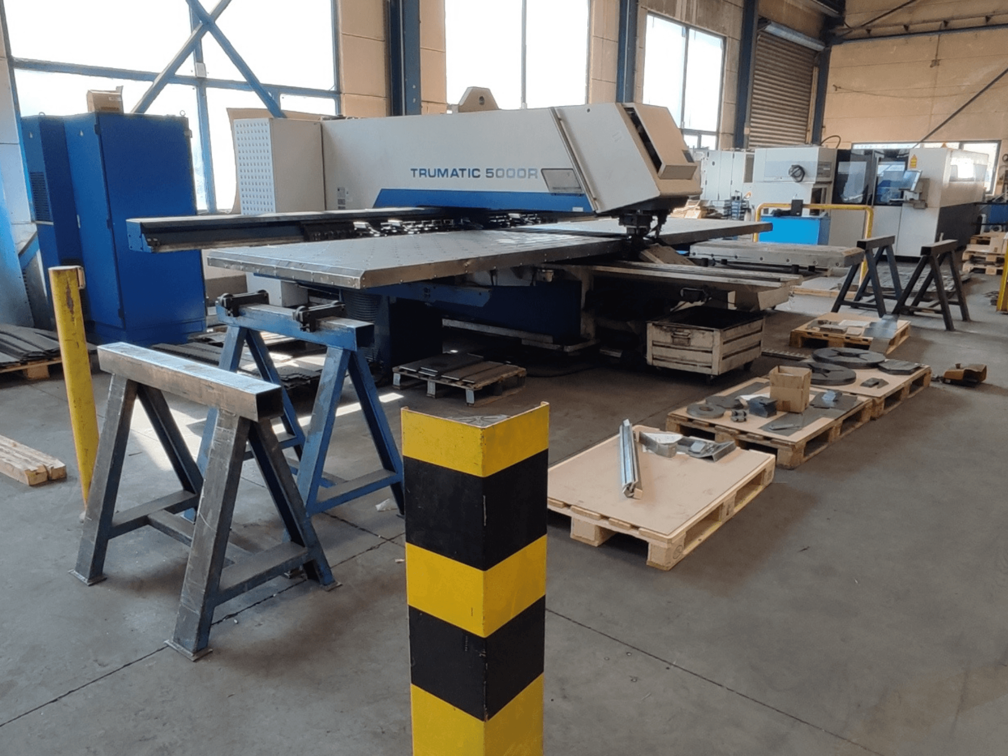 Poinçonneuse CNC TRUMPF Trumatic 5000 R dans un atelier, vue de profil avec des outils et des matériaux posés sur des palettes à proximité.