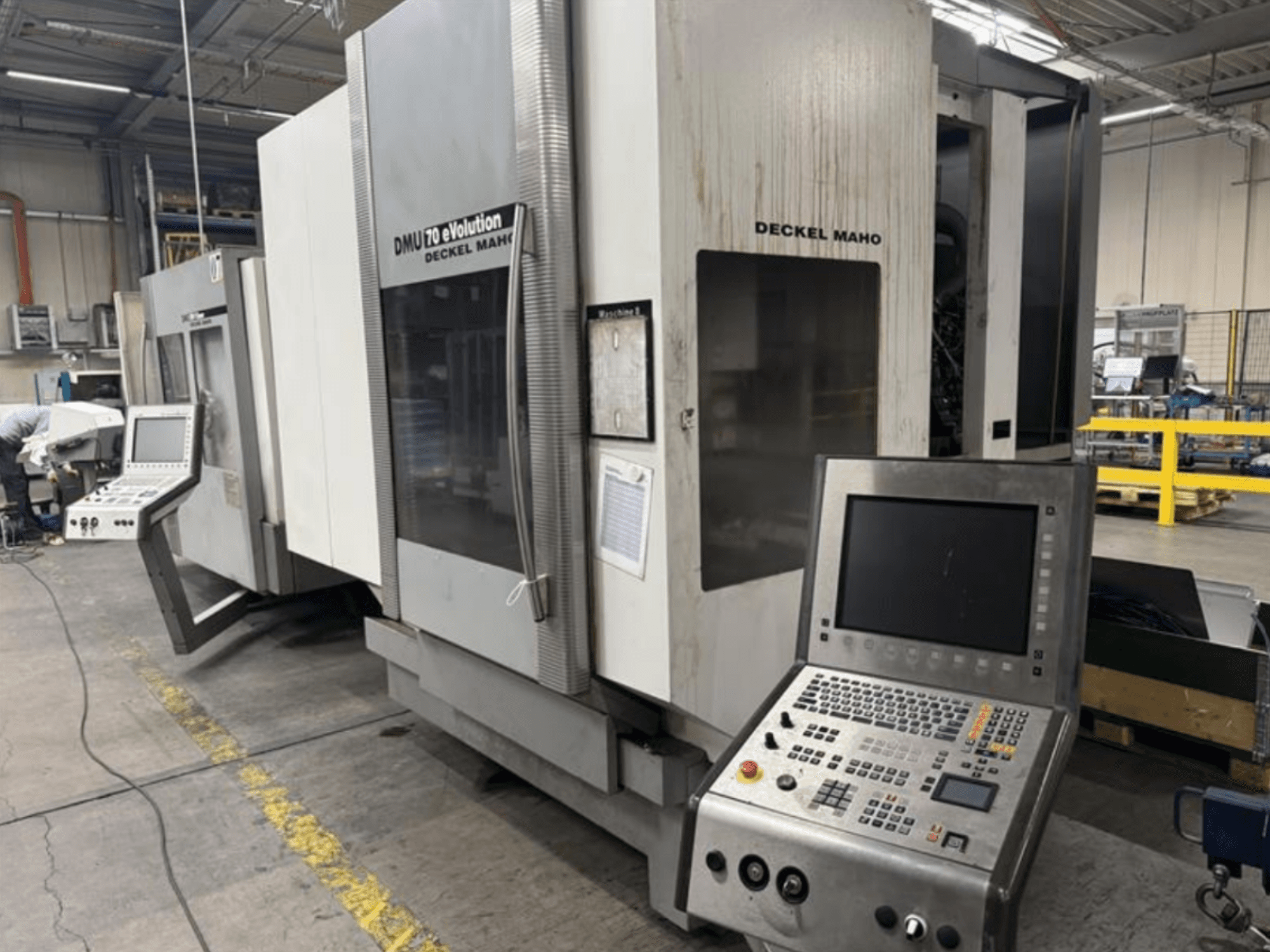 Vue de face d'une machine CNC Deckel Maho DMU 70 eVOLUTION avec panneau de commande, située dans une usine de fabrication.
