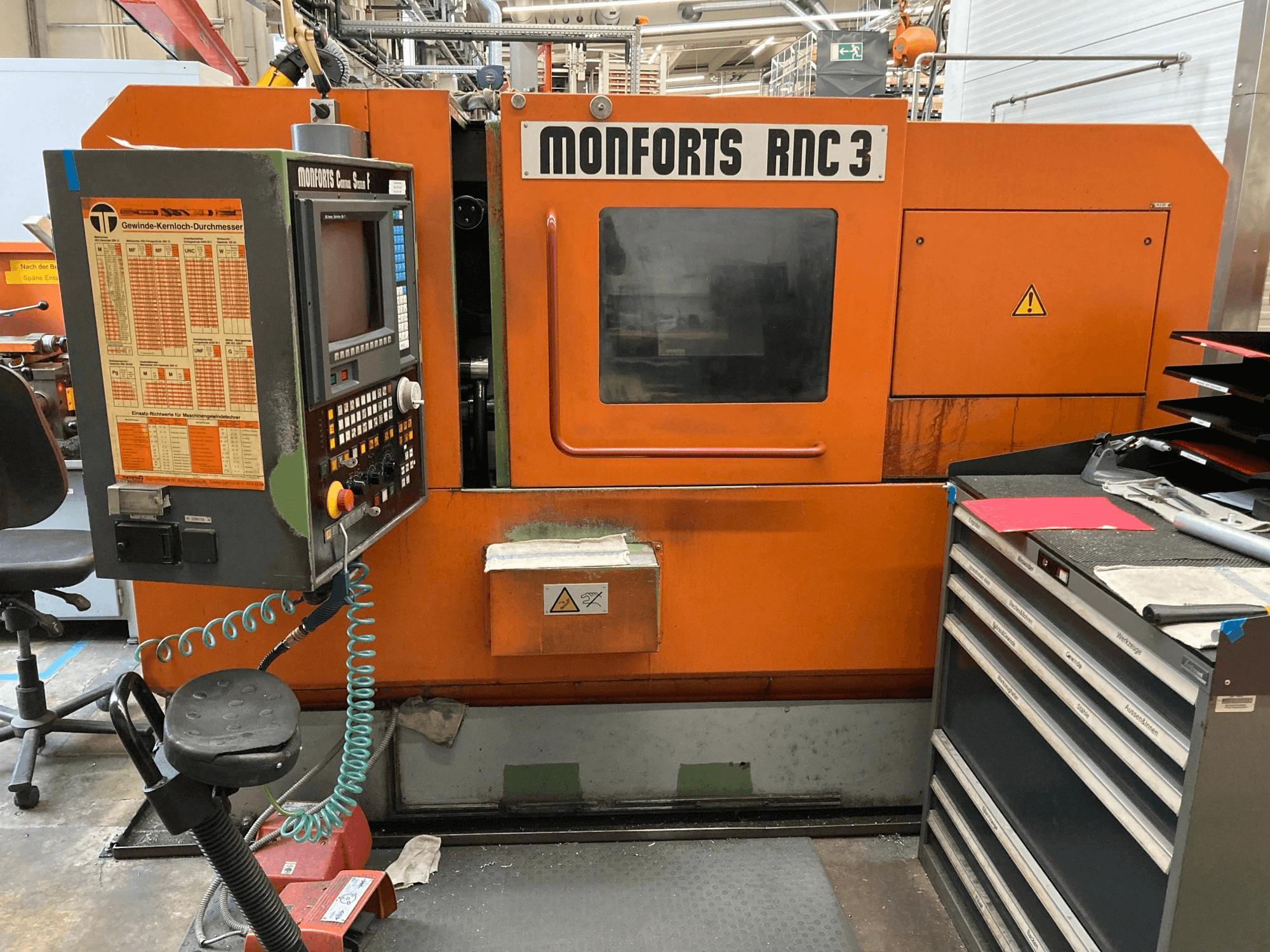 Machine Monforts RNC 3 en orange et noir, vue de face, comportant un panneau de commande et divers tiroirs à outils.