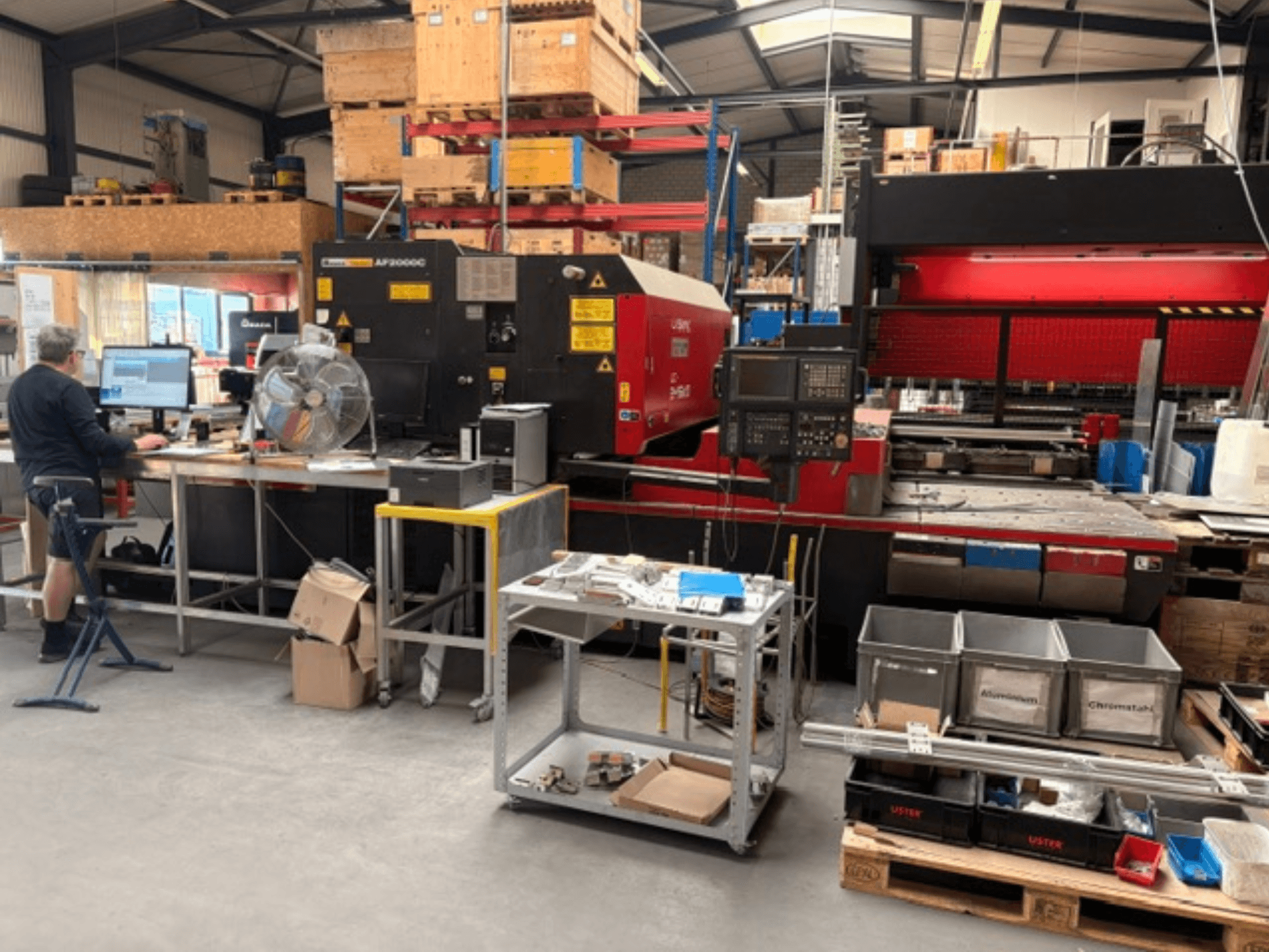 Machine de découpe laser CO₂ AMADA LC-2415αIII dans un atelier, avec un poste de travail informatique visible au premier plan.