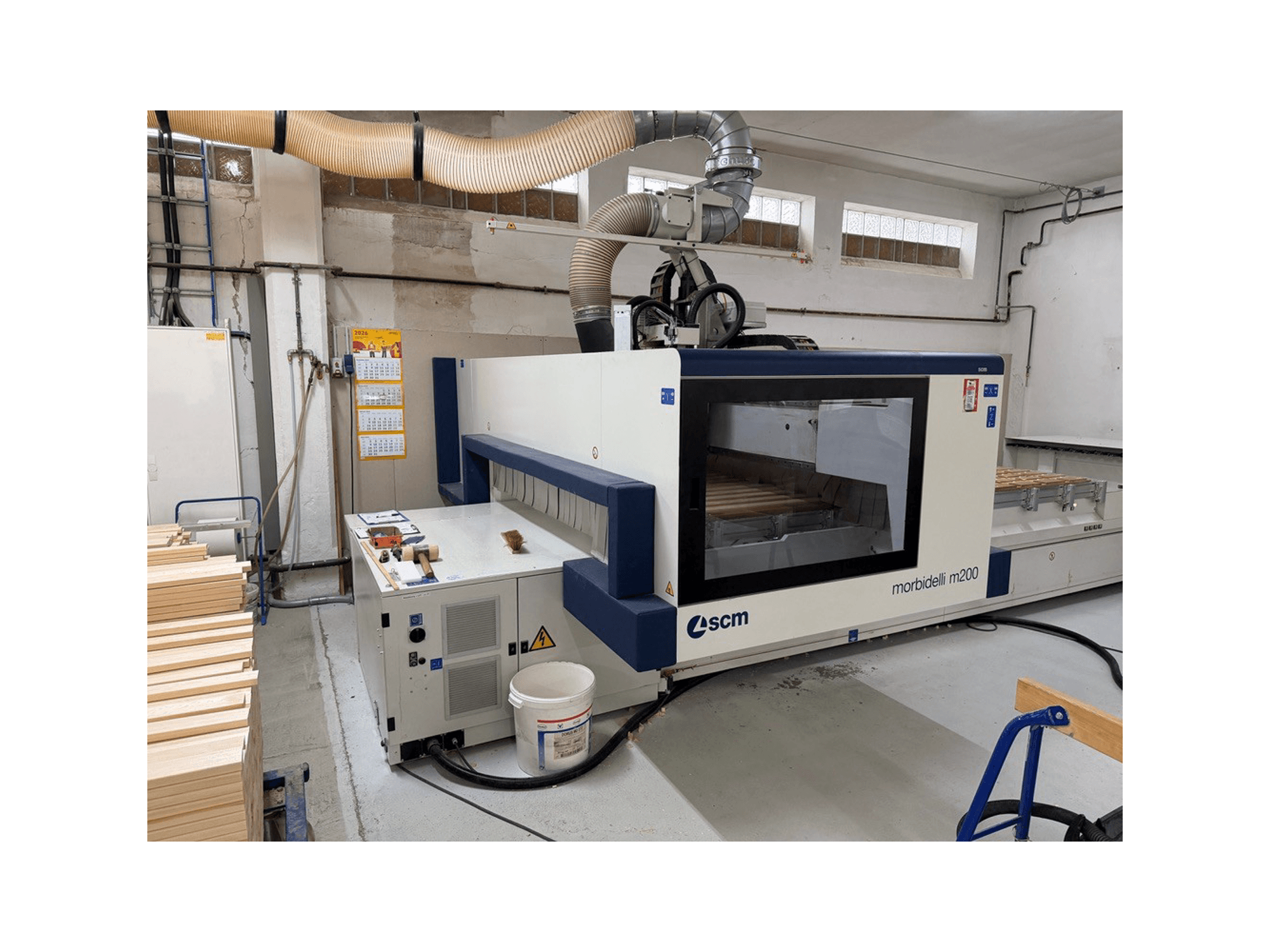 Centre d'usinage CNC SCM Morbidelli M200F dans un atelier, vue de profil avec des pièces de bois sur la table et un système de ventilation au-dessus.