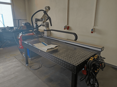 Cellule de soudage par impulsions FRONIUS TransSteel 4000 vue de profil, comprenant un bras robotisé, un espace de travail et des raccordements électriques.