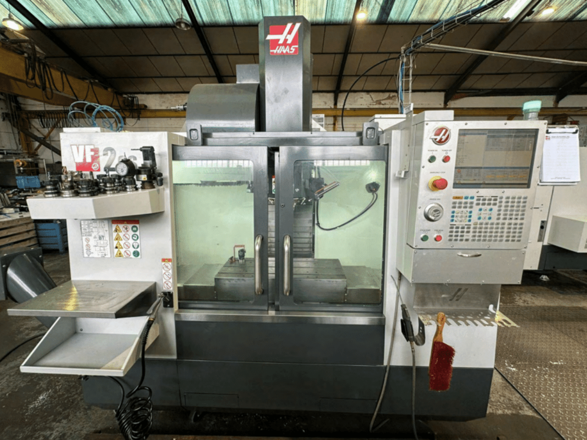 Vue avant de la machine CNC Haas VF2, montrant le panneau de commande, les porte-outils et l'espace de travail avec des étiquettes de sécurité et une brosse.