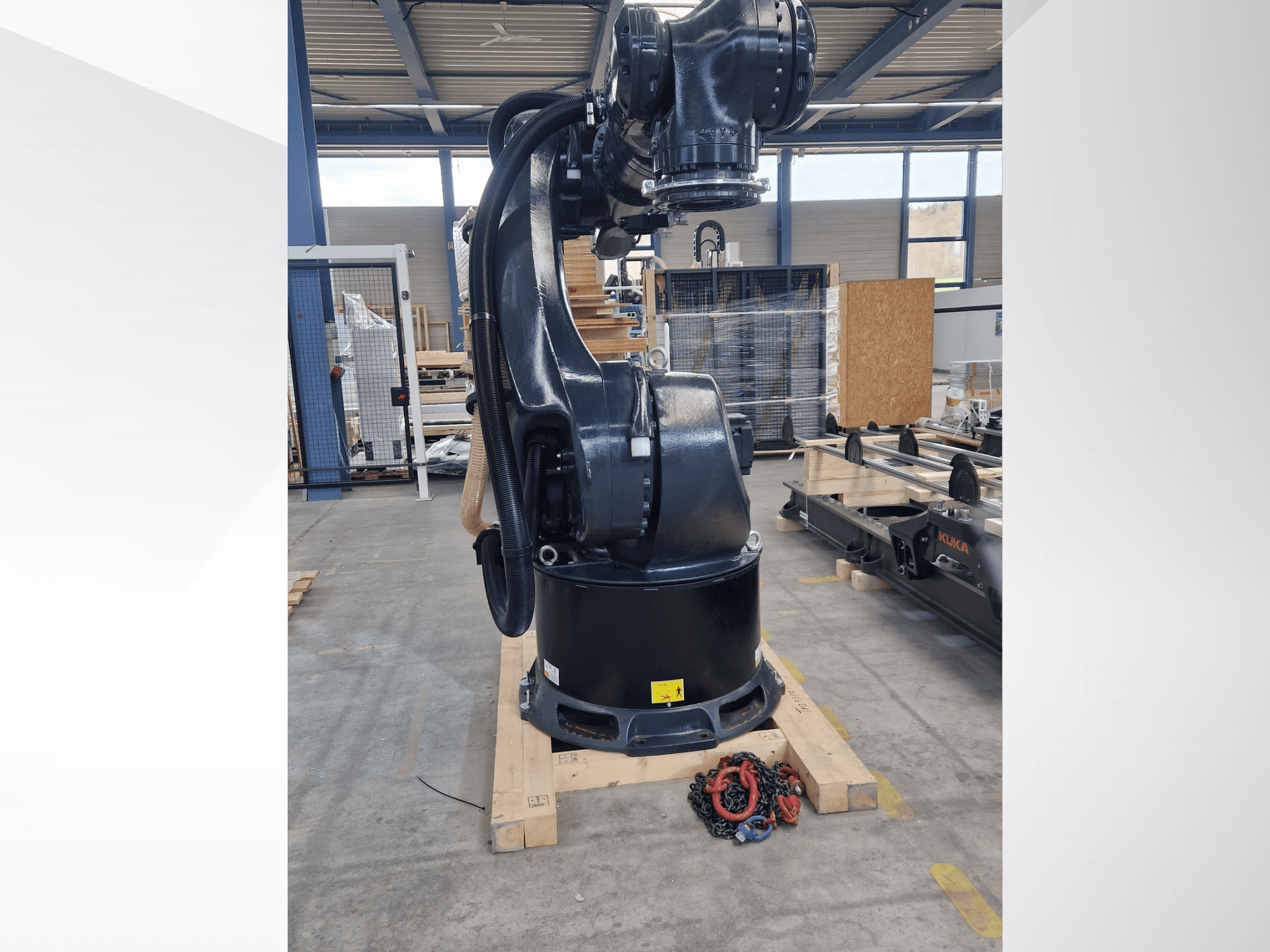 Bras robotisé KUKA KR 340 R3330 dans un entrepôt, vue de profil montrant sa base et ses câbles, avec des chaînes au premier plan.