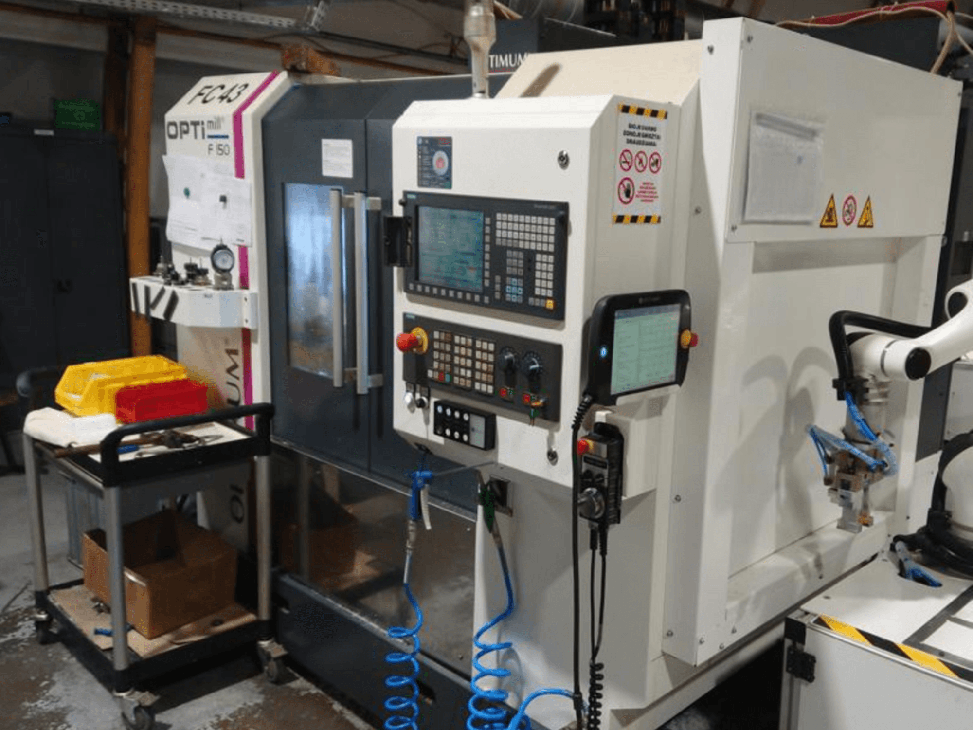 Panneau de commande d'une fraiseuse OPTIMUM F50 avec des tuyaux, des outils et un bras robotisé visibles dans un atelier.