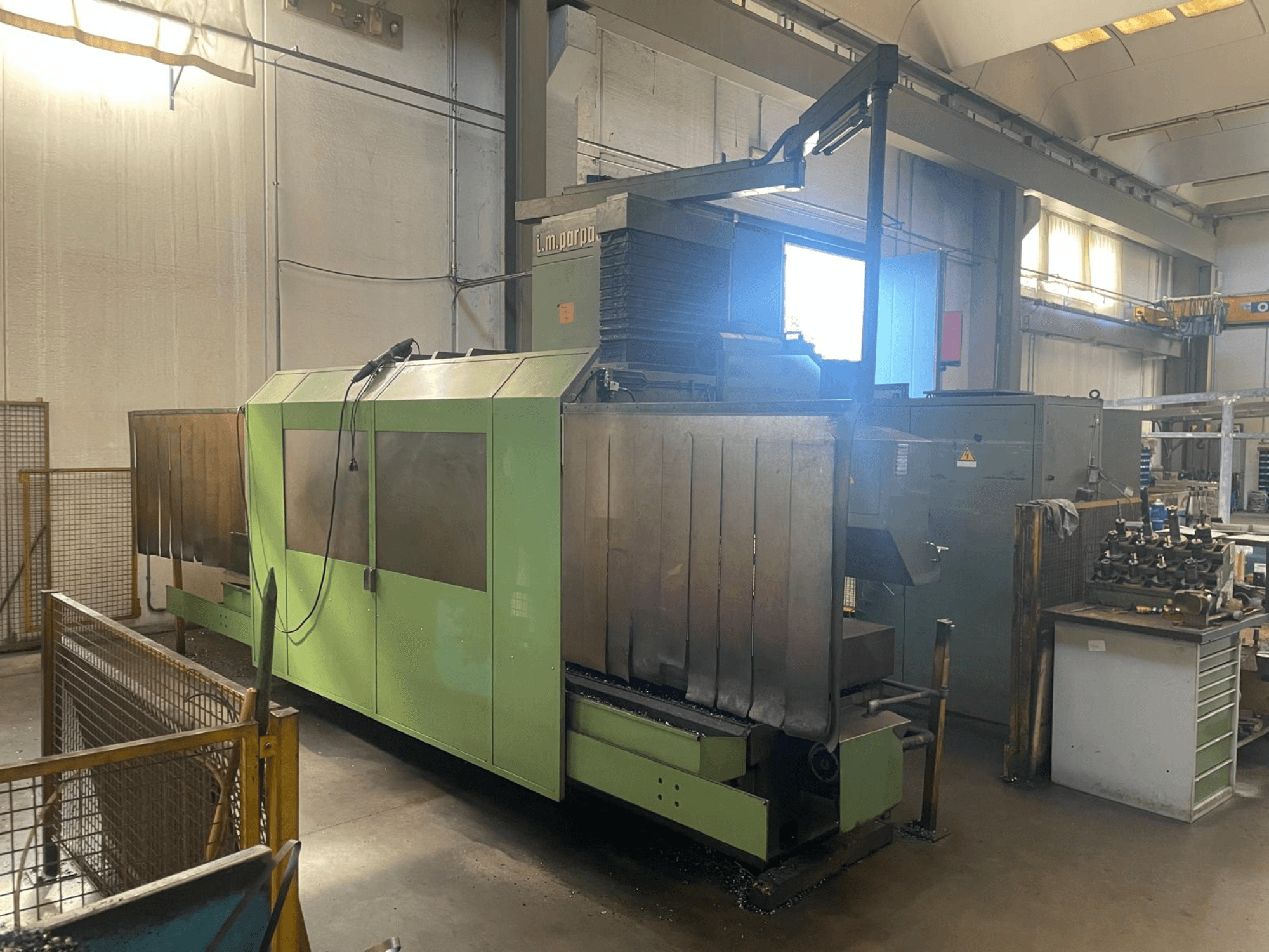 Machine industrielle verte avec des composants métalliques visibles, vue de face ; elle comporte un panneau de commande, des avertissements de sécurité et un poste de travail à proximité.