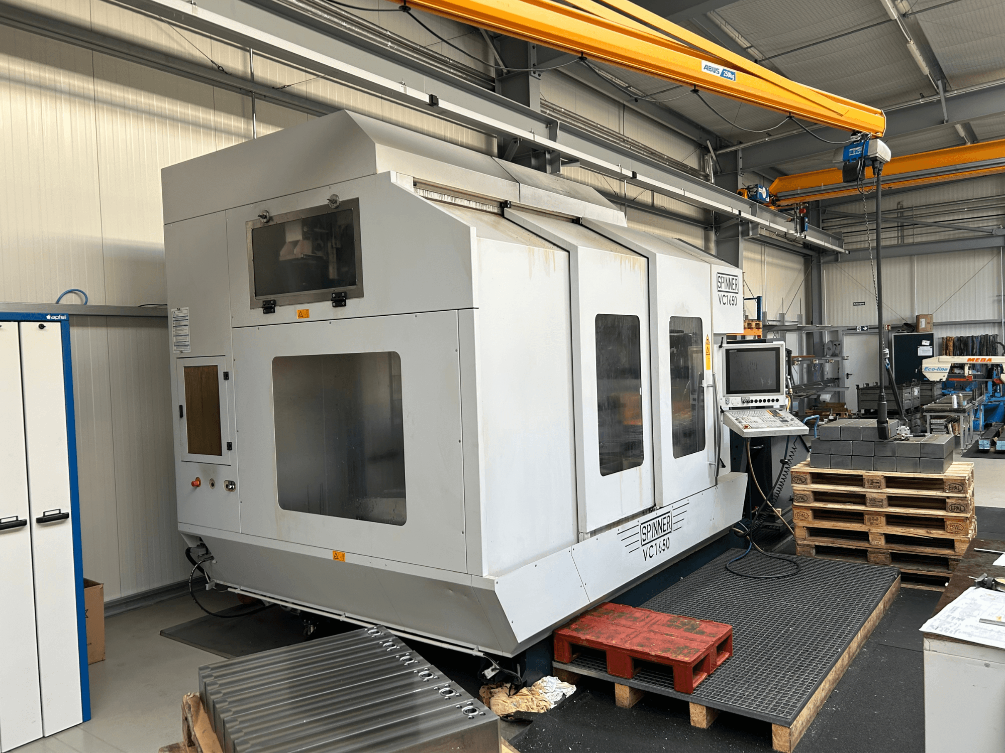 Machine CNC Spinner VC 1650, vue de face, avec panneau de commande, grandes fenêtres et palettes empilées à proximité dans un atelier.