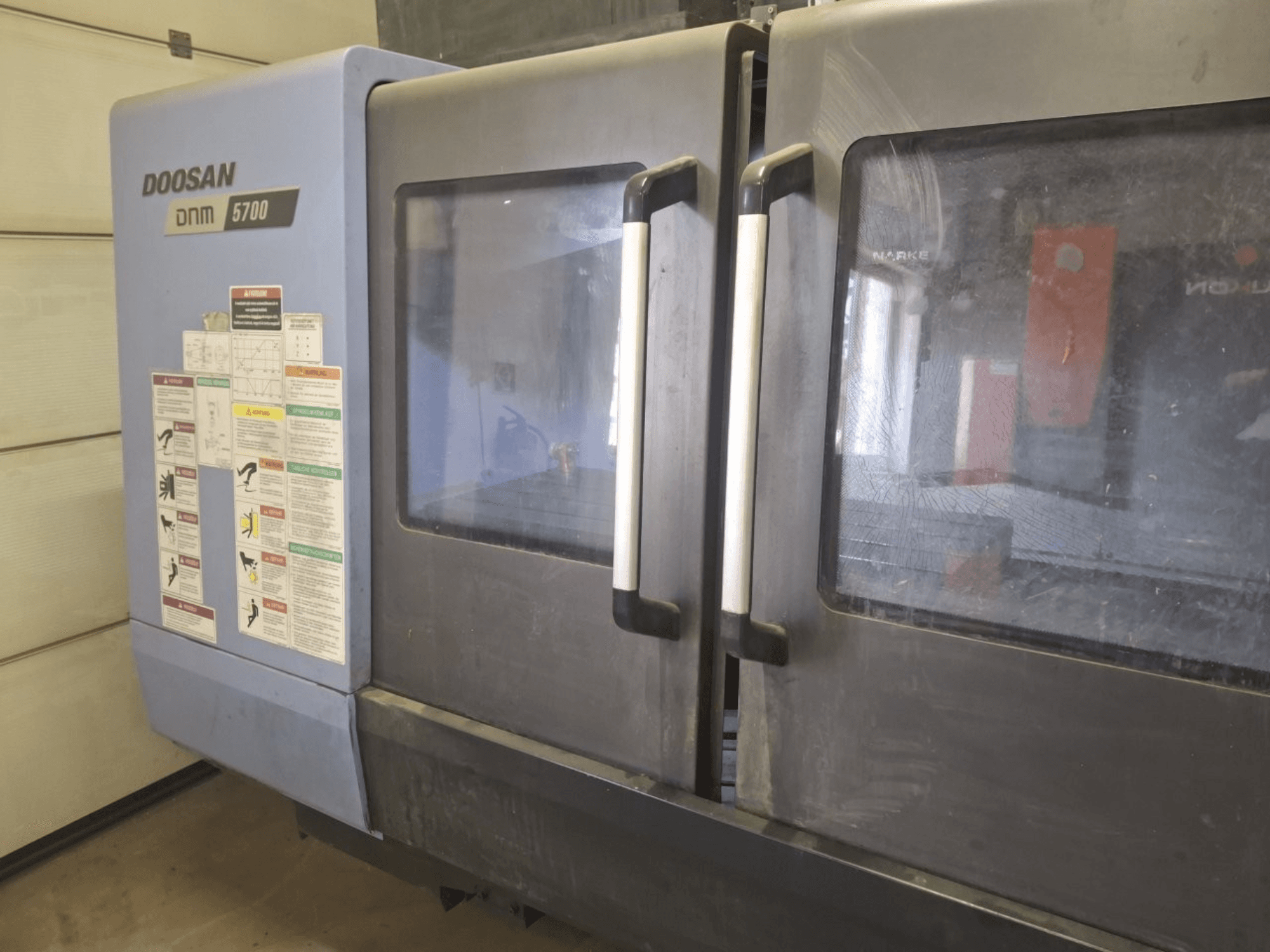 Vue de face d'un centre d'usinage vertical DOOSAN DNM 5700, présentant des portes doubles et des panneaux de commande avec des directives opérationnelles.