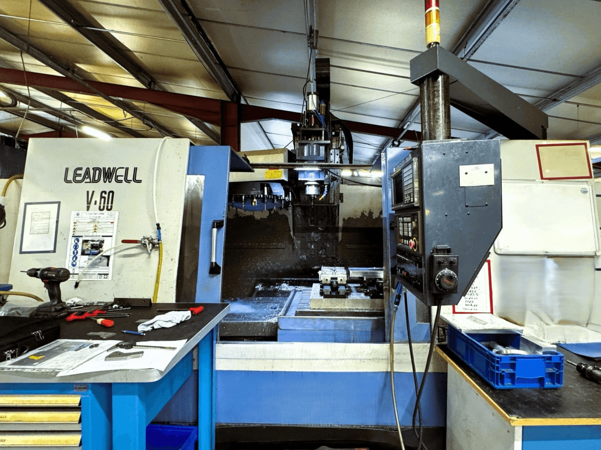 Vue de profil du centre d'usinage vertical Leadwell V-60, mettant en évidence le panneau de commande et la zone de travail avec les outils et les pièces.