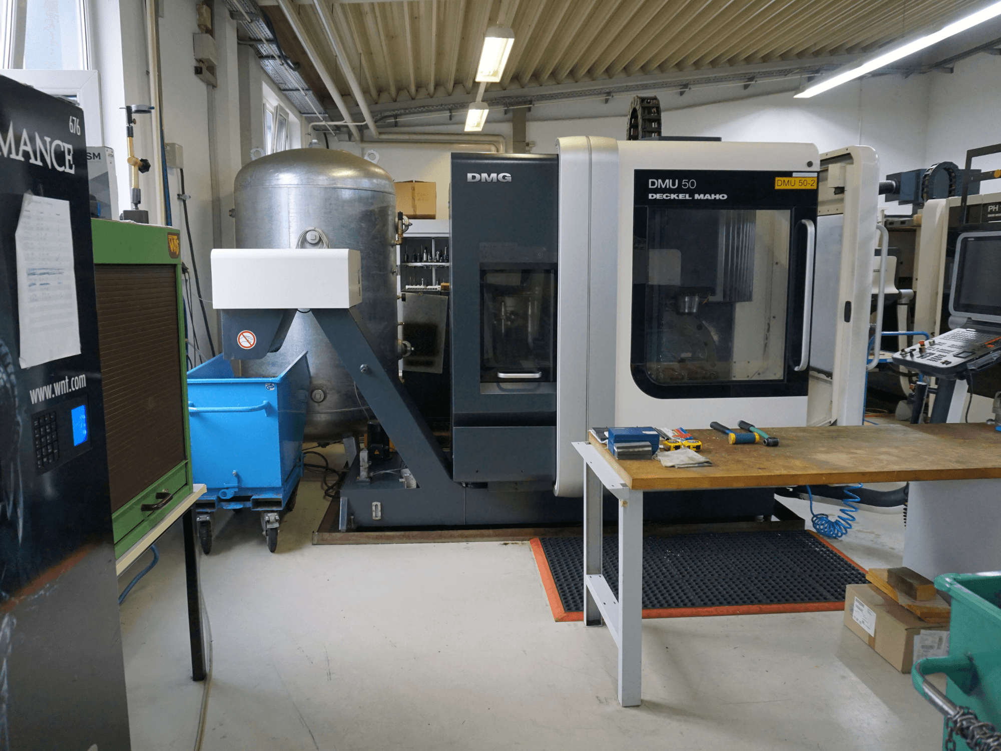 Fraiseuse DMG DMU 50 dans un atelier, vue de face, avec des outils sur une table en bois et une poubelle à roues bleues à proximité.