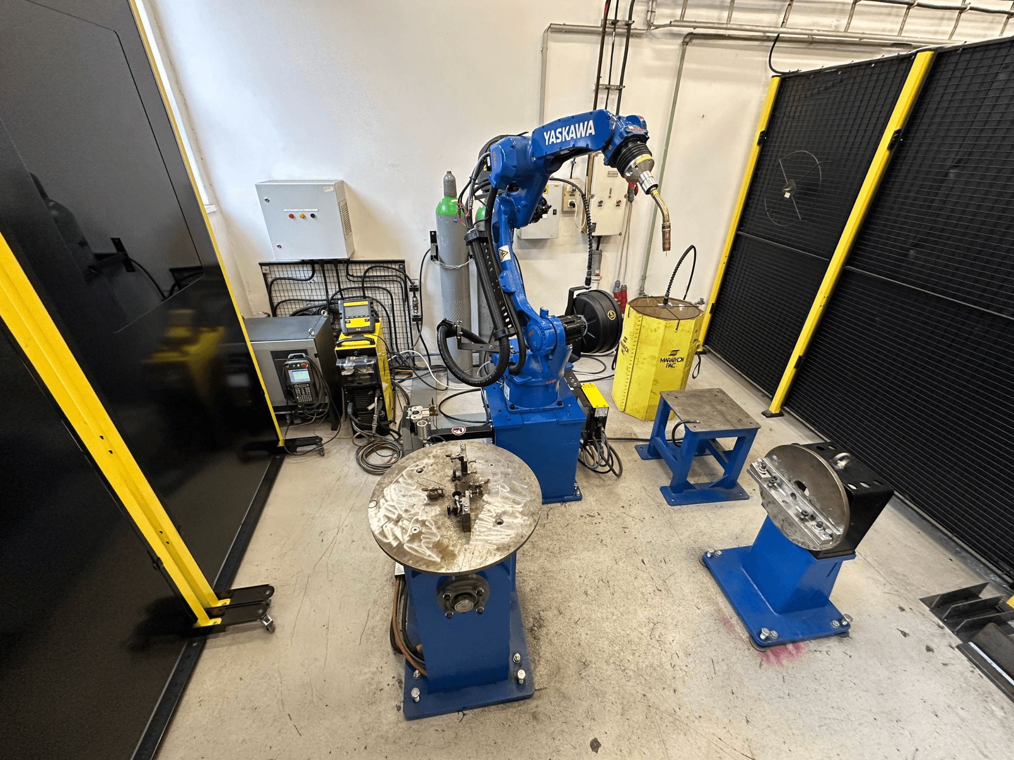 Bras robotique Yaskawa en bleu avec deux tables de travail, du matériel de soudage et des barrières de sécurité ; vue de face d'un espace de travail industriel.