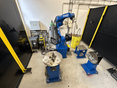 Bras robotique Yaskawa en bleu avec deux tables de travail, du matériel de soudage et des barrières de sécurité ; vue de face d'un espace de travail industriel.