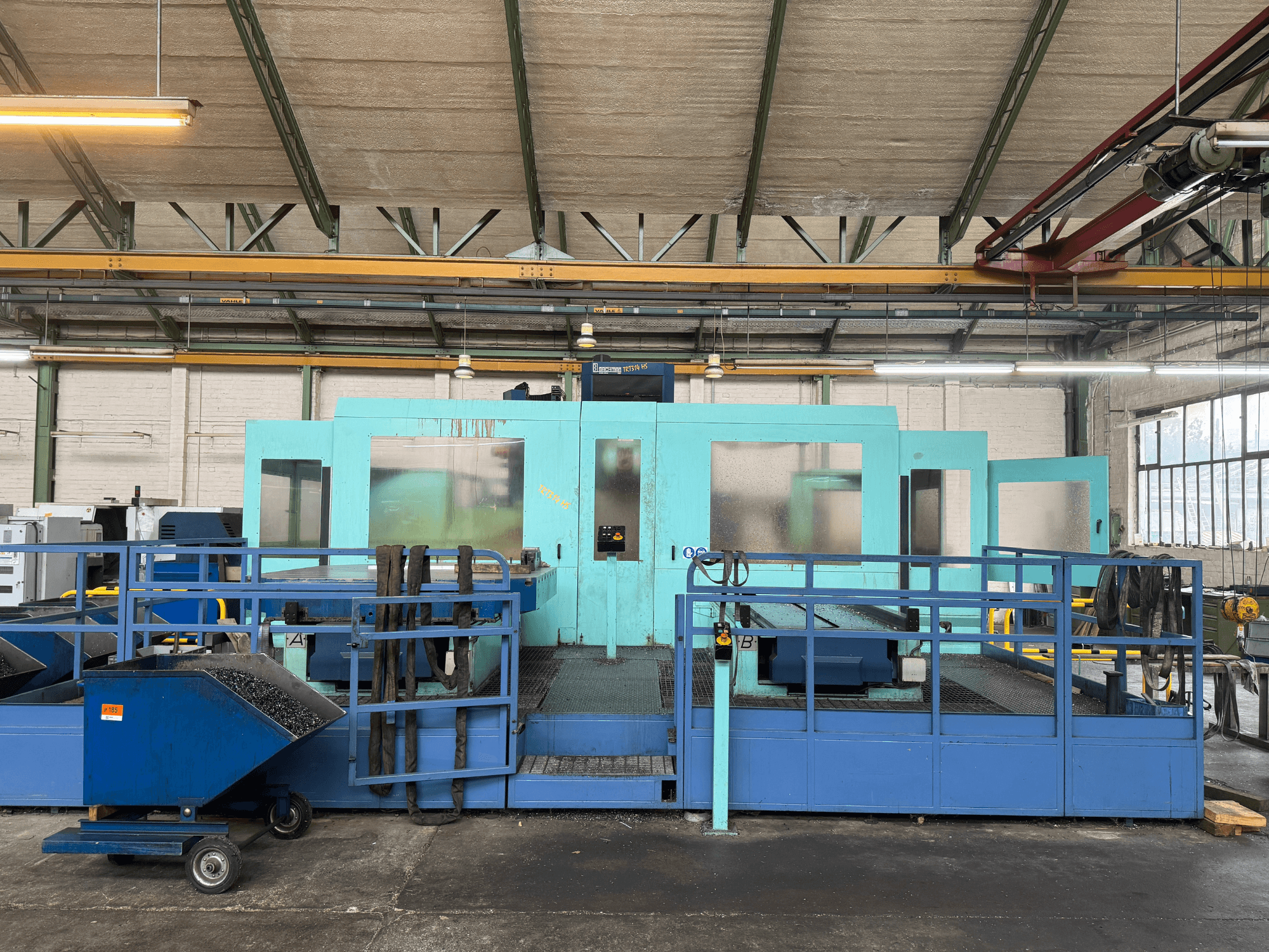 Machine CNC industrielle bleue avec panneaux de protection, vue de face, comprenant un rangement pour les outils et un chariot de collecte des déchets sur le côté.