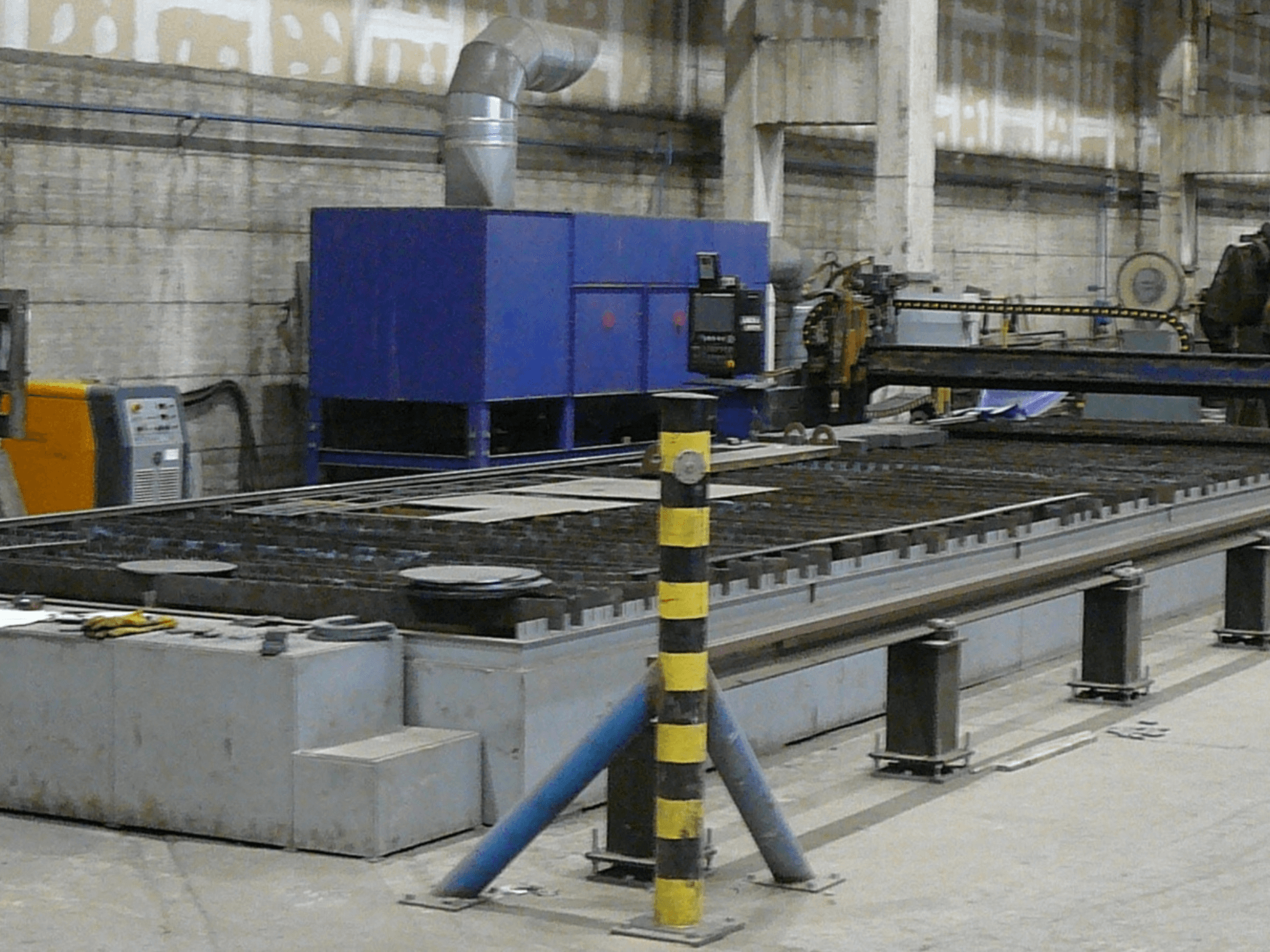 Installation d'une machine industrielle de découpe des métaux, vue de côté, comprenant un boîtier de commande bleu, des bandes de sécurité et une surface de travail en métal.