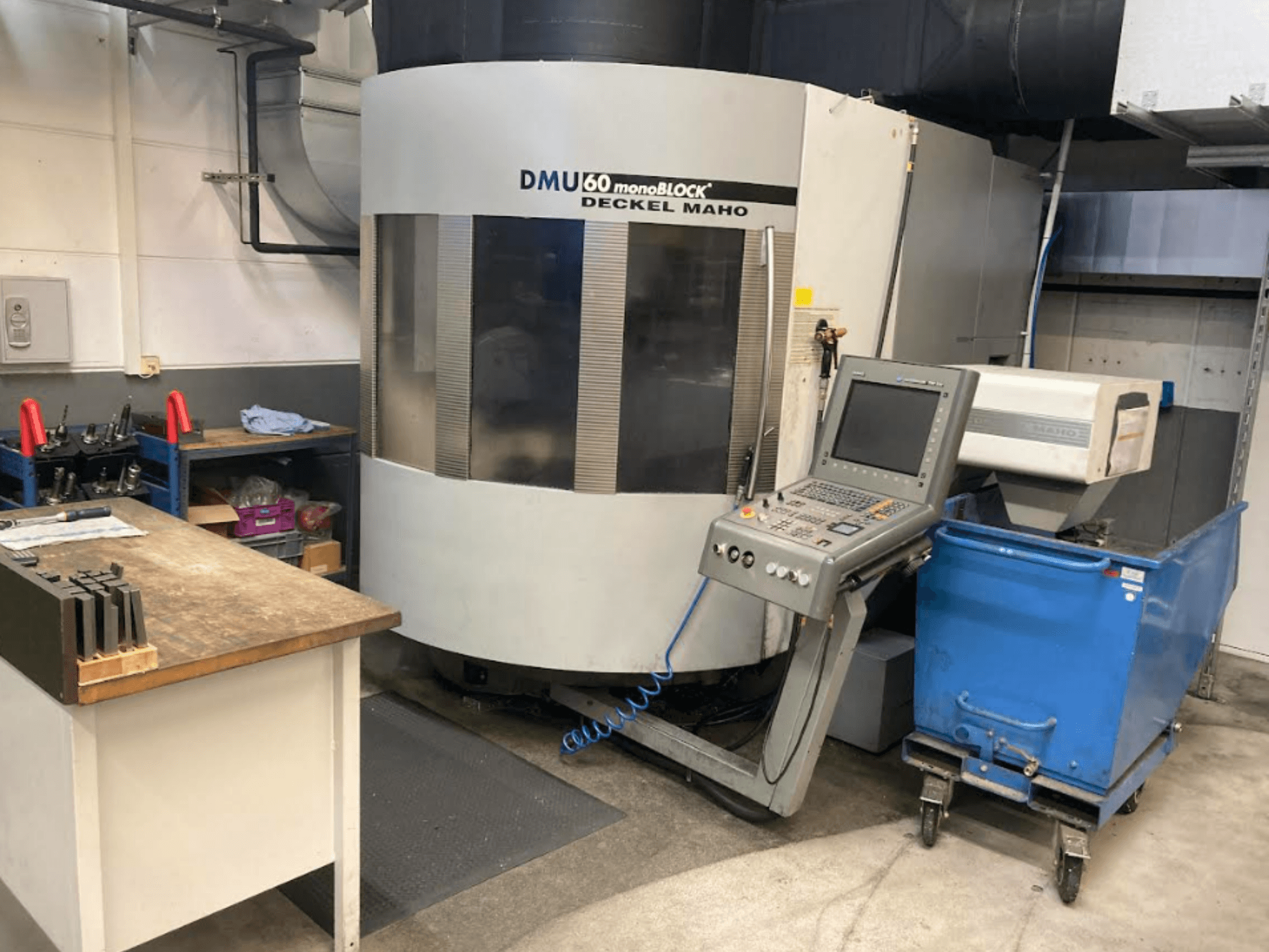 Centre d'usinage vertical DMG MORI DMU 60 Monoblock, vue de face avec le panneau de commande, l'espace de travail et le chariot d'outils bleu visibles.